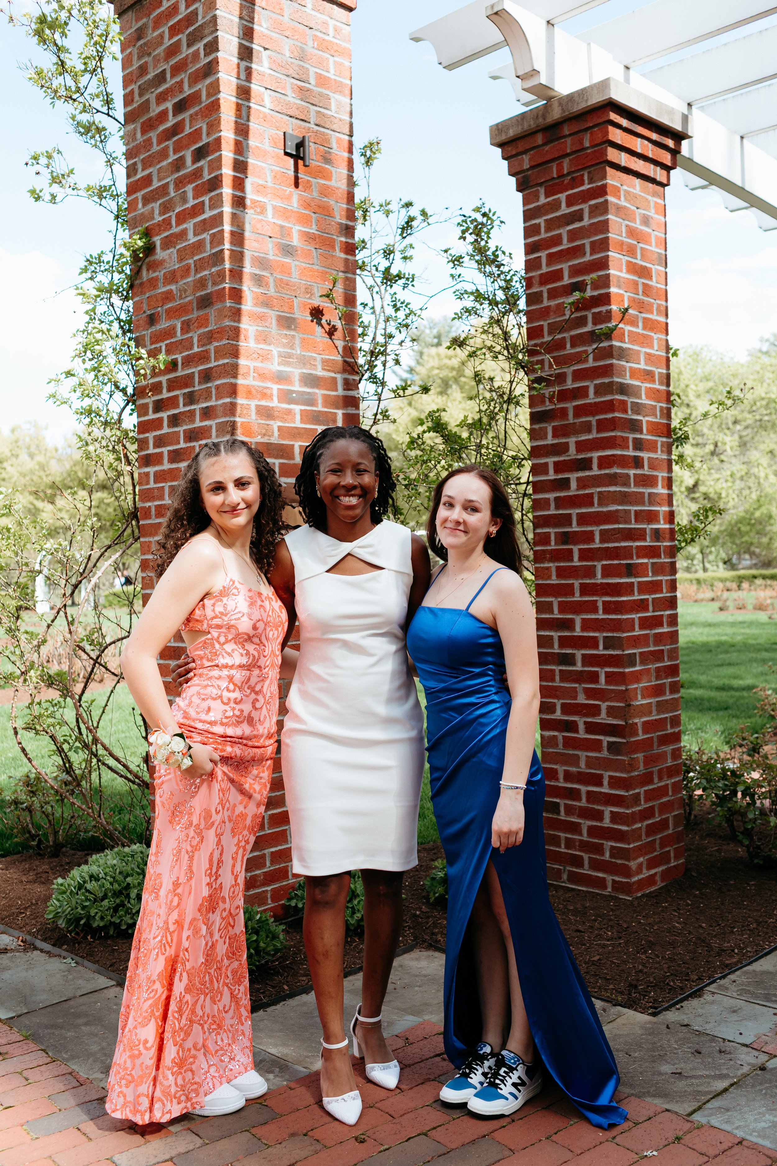 Three young women posing in formal dresses outside near brick columns and greenery.