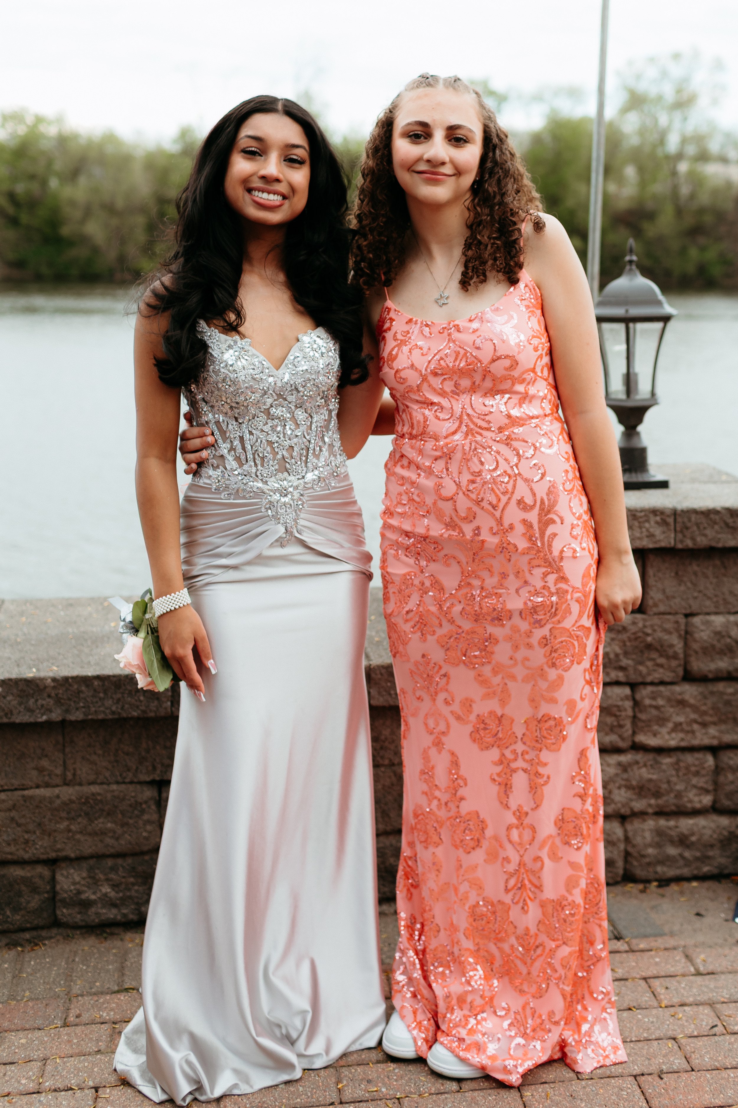 Two people wearing formal gowns, standing outdoors by a stone wall with a body of water in the background.