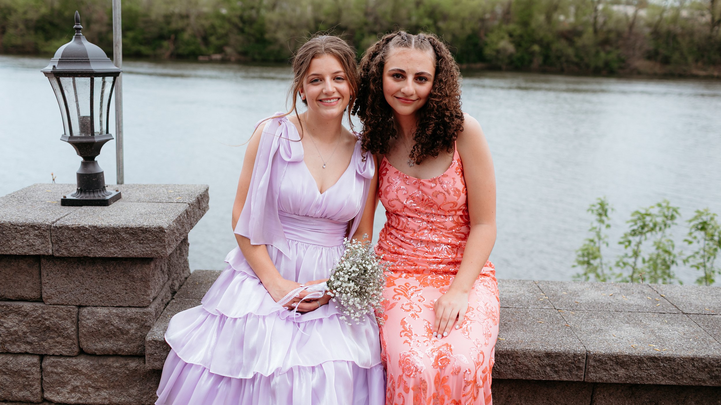 Two young women in formal dresses sitting by a river on a stone wall, one in lavender and one in pink, with a vintage-style lamppost nearby.