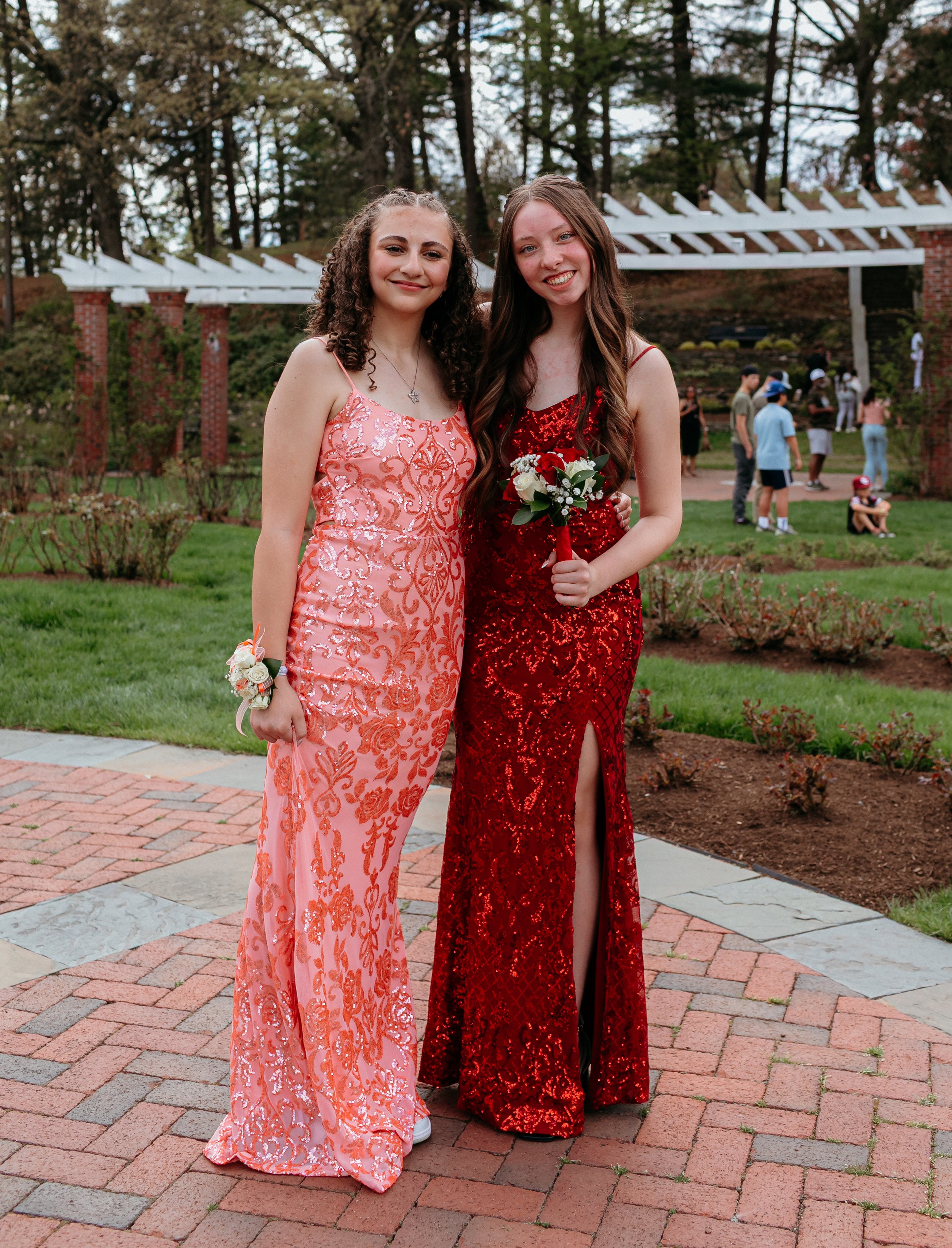 Two women wearing evening gowns, one in pink and one in red, standing on a brick walkway in a garden. The woman in red is holding a small bouquet. Trees and other people are in the background.