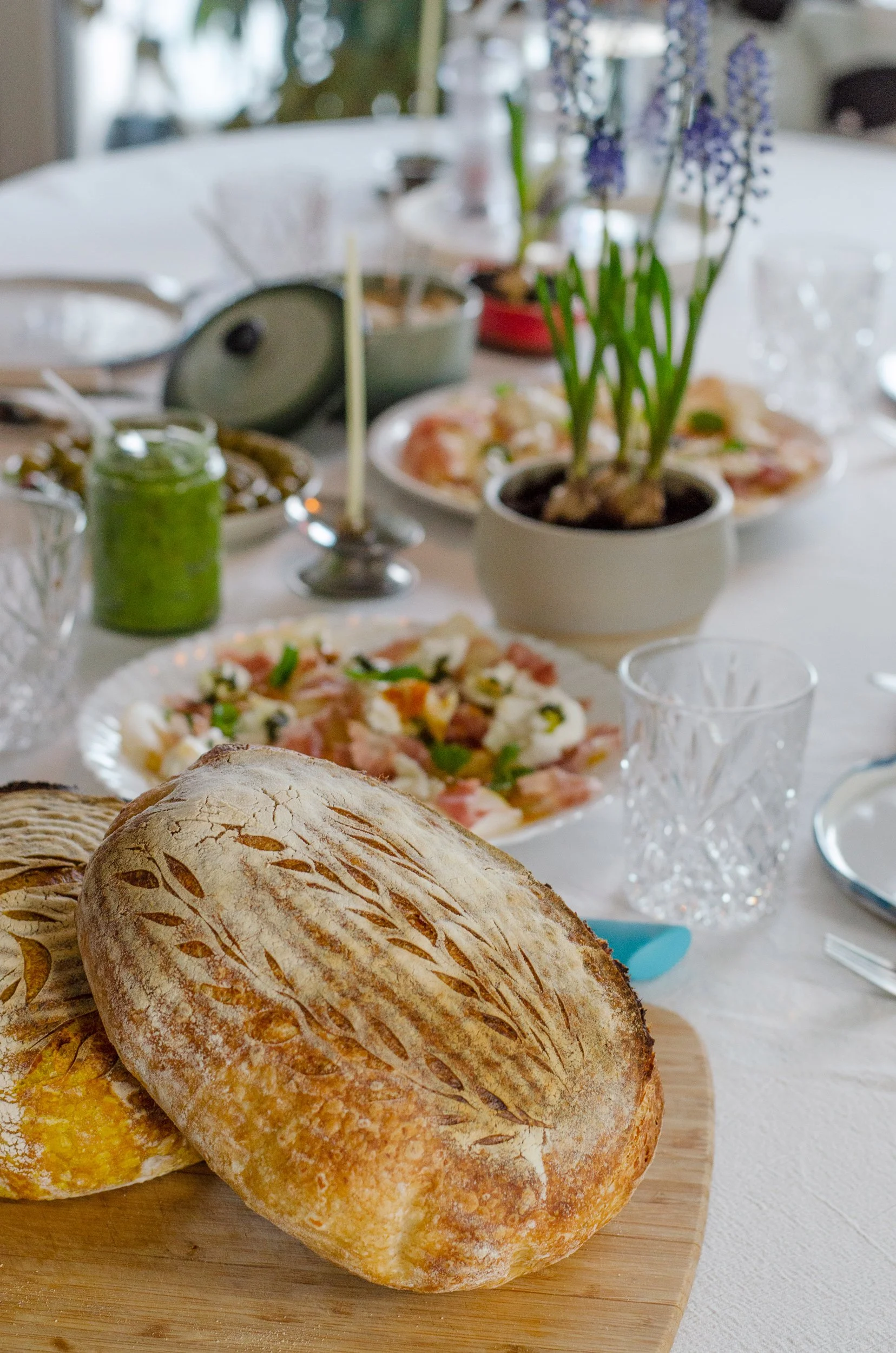 Freshly baked sourdough bread loaves on a wooden cutting board in the foreground with a table set with various dishes, a plant, a small candle, and glasses in the background.