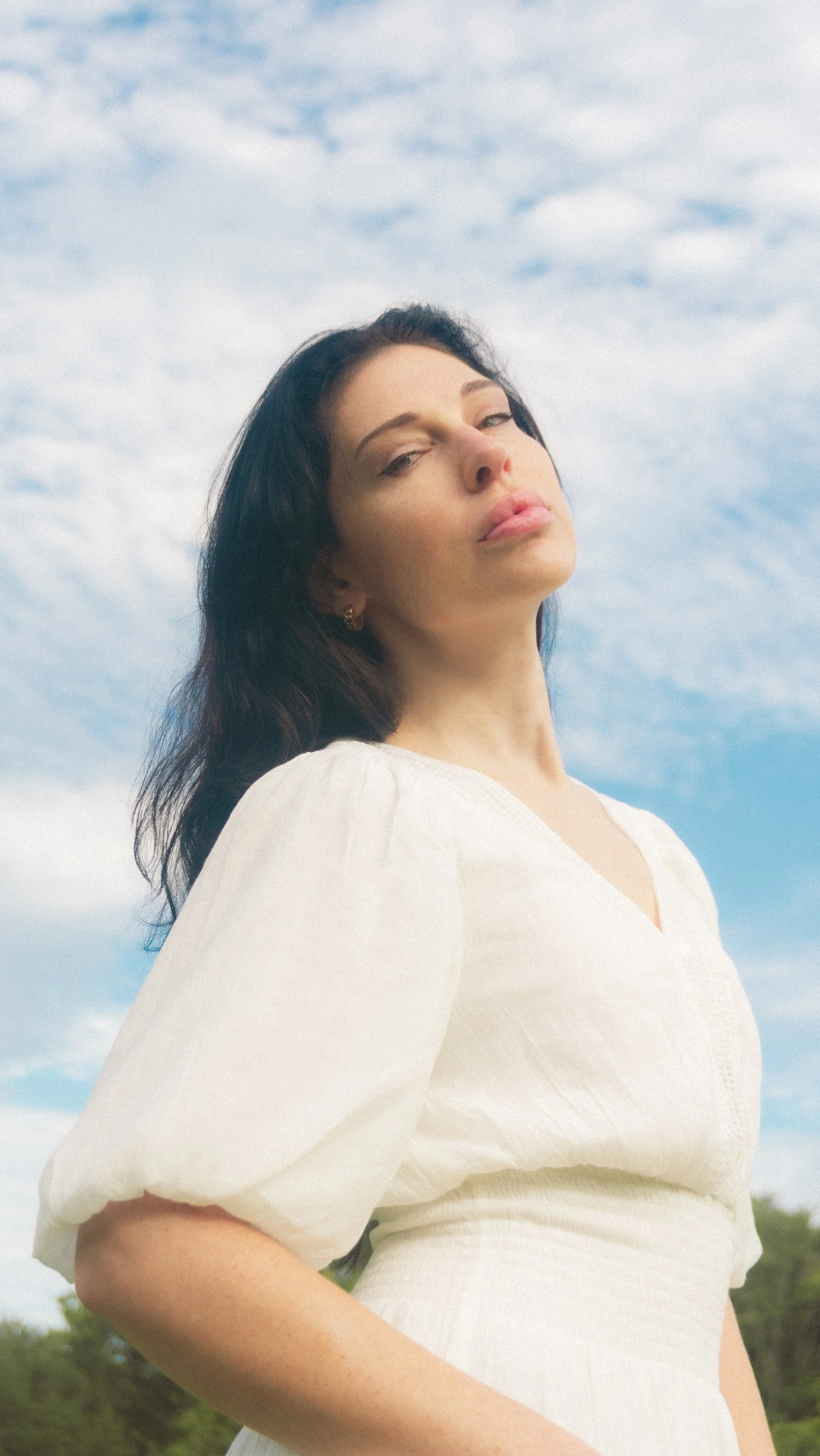 A woman in a white dress with a blue sky background looks at the camera