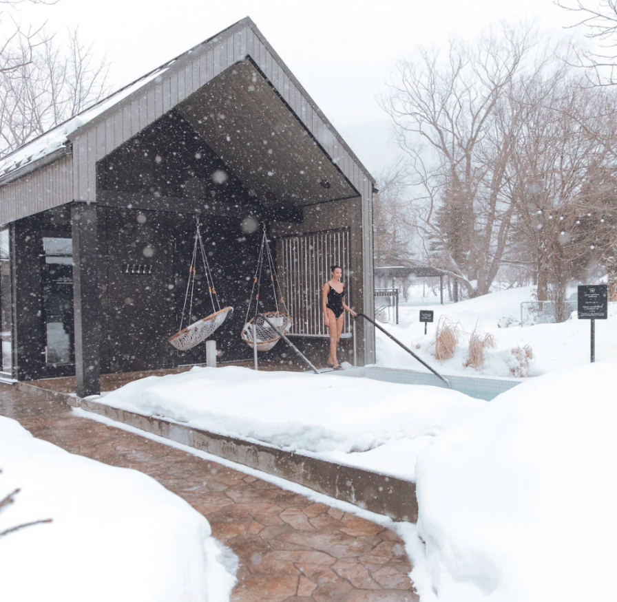 A woman in a black swimsuit climbing out of a hot tub with trees and additional hot tubs in the background.