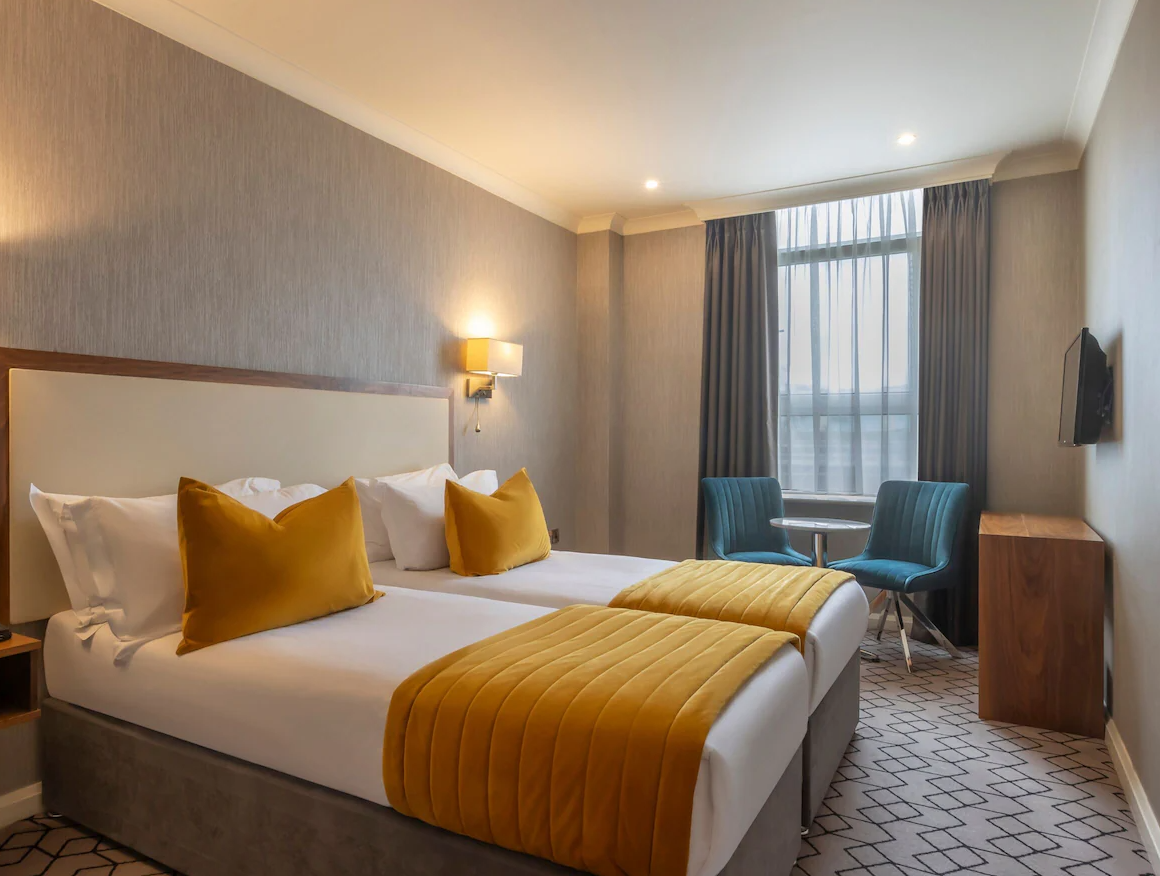 A hotel room with two beds, mustard yellow pillows and bedspreads, blue chairs, a window with curtains, and a wall-mounted TV.