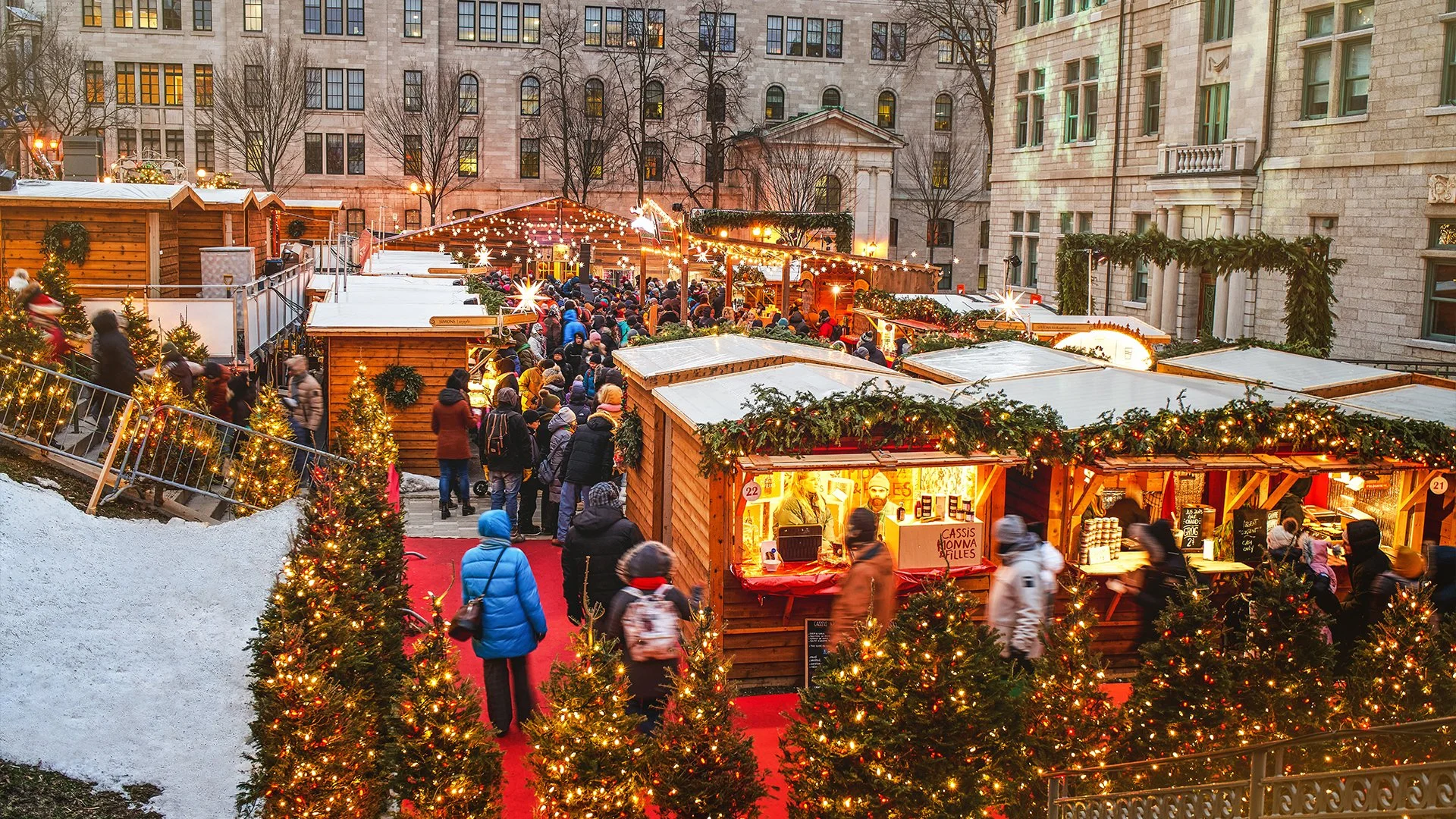 Christmas market in a city square at night with a decorated Christmas tree, holiday stalls, and crowds of people under falling snow.