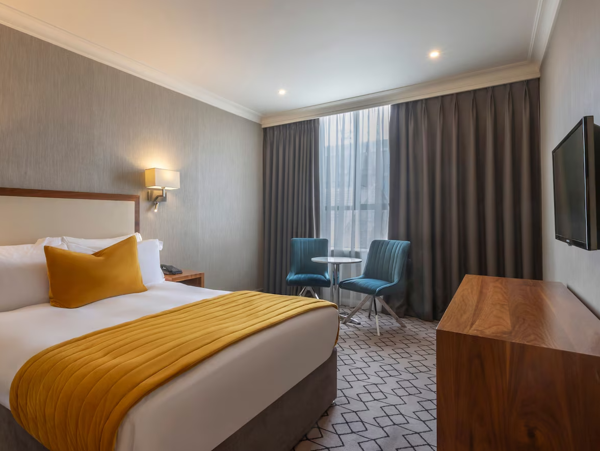 Hotel room with a bed having yellow bedding and pillows, a side table, two blue chairs with a small round table near a large window with curtains, a wall-mounted TV, and a patterned carpet.