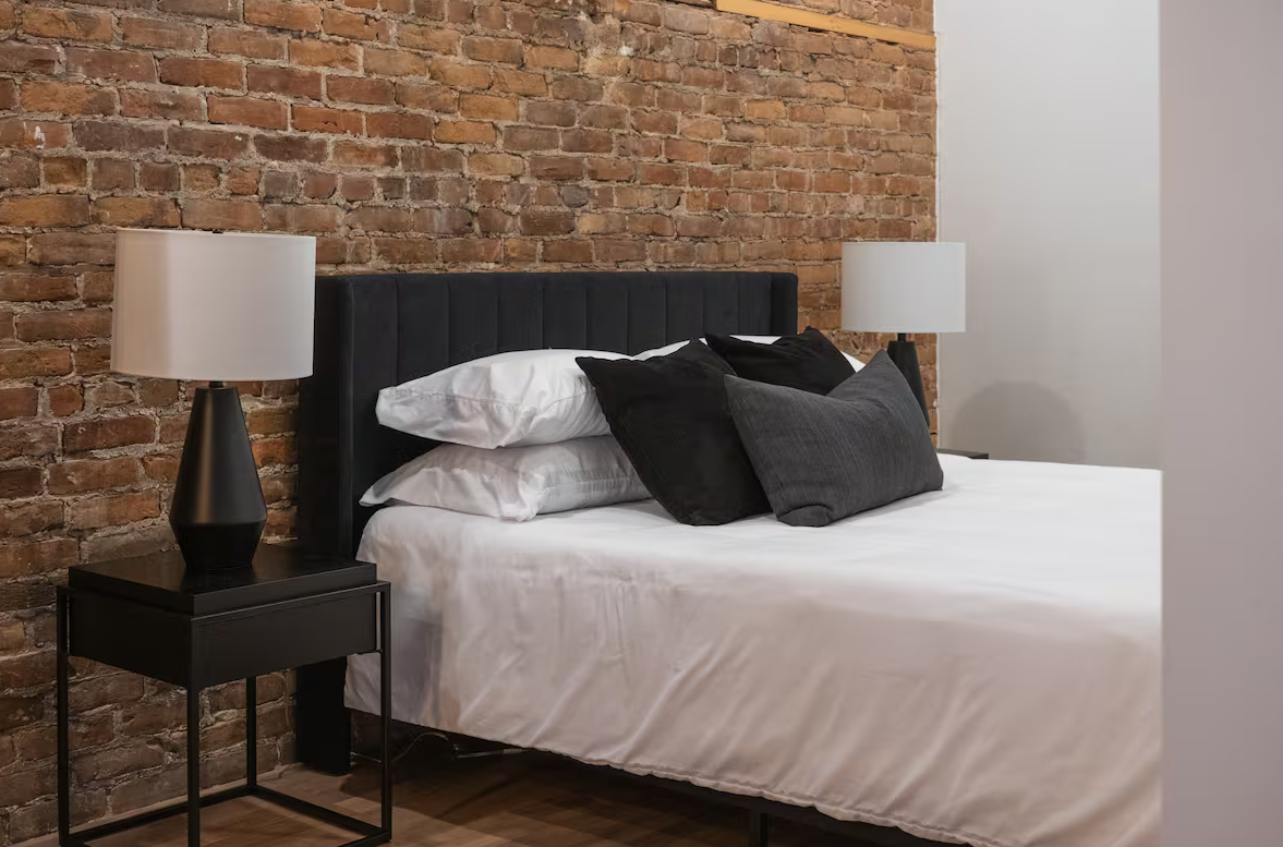 A modern bedroom with a bed that has white sheets and black and gray pillows, a black headboard, two black nightstands with white lamps, and an exposed brick wall.