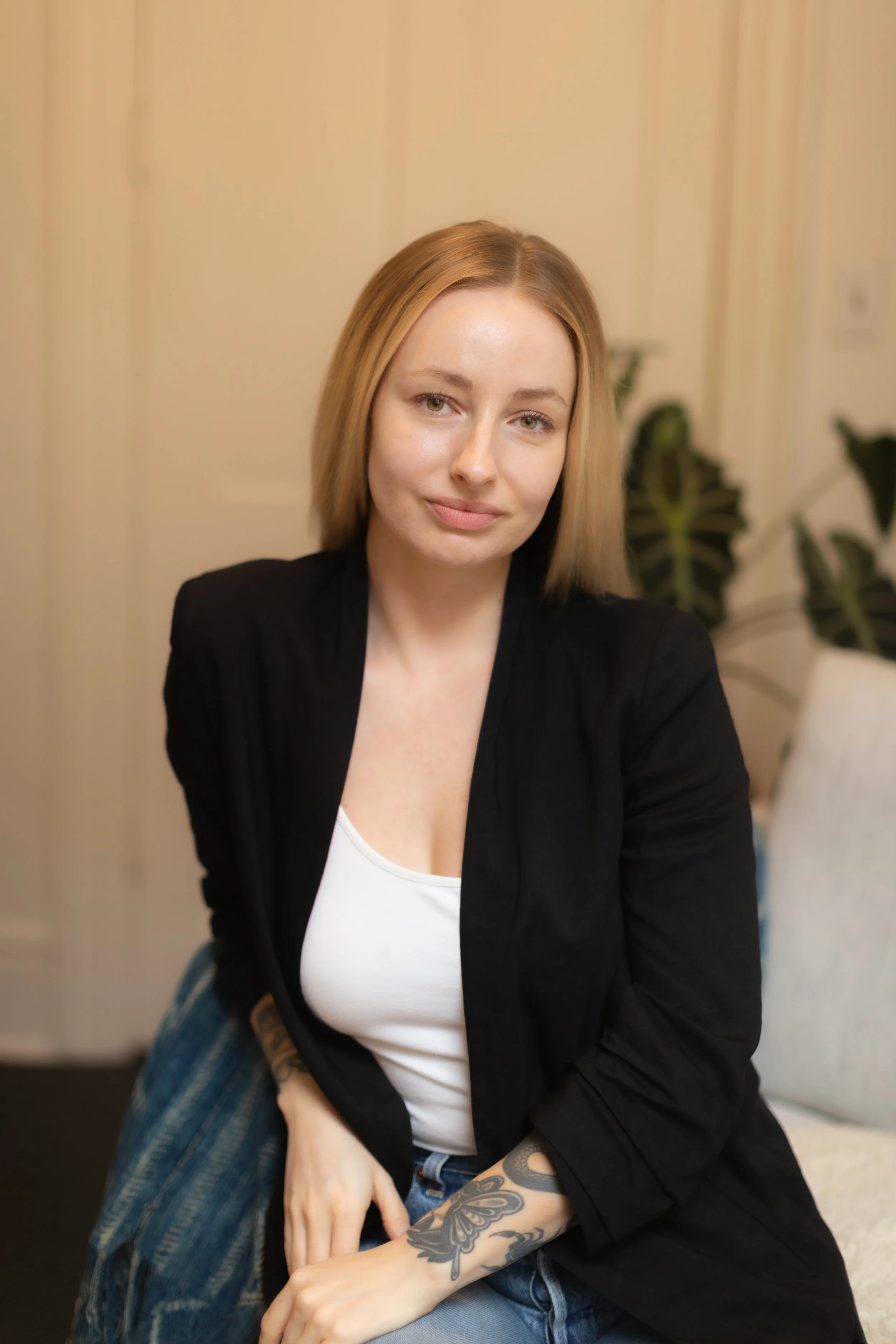 A person with medium-length blonde hair, wearing a black blazer over a white top, sitting on a blue chair. The individual has tattoos on their forearms and is in a room with light-colored walls and a green plant in the background.