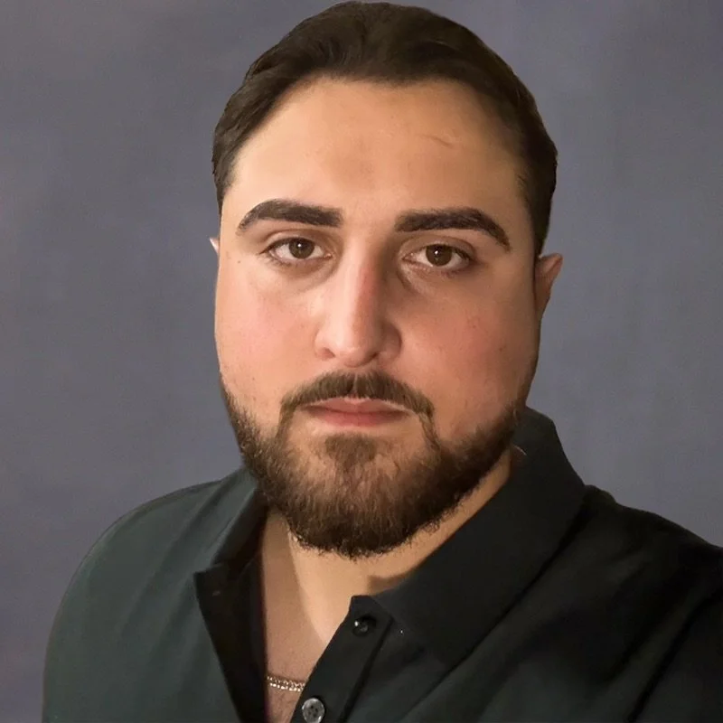 A close-up portrait of a man with dark hair, a beard, and mustache, wearing a black collared shirt and a silver chain necklace, against a dark gray background.
