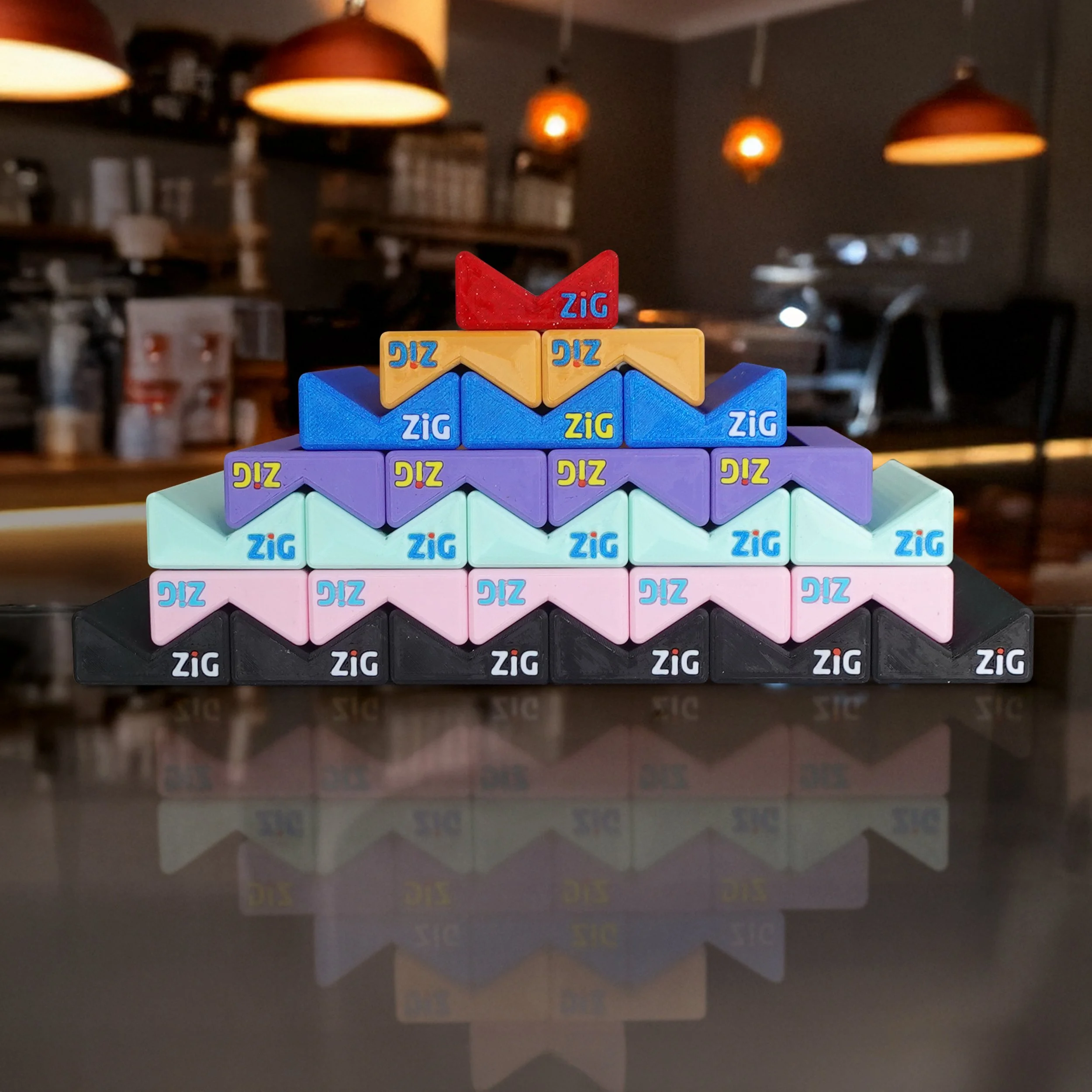 Stack of colourful  ZigaJig King Slim Rolling Rack arranged in a pyramid shape on a reflective surface in a café setting.