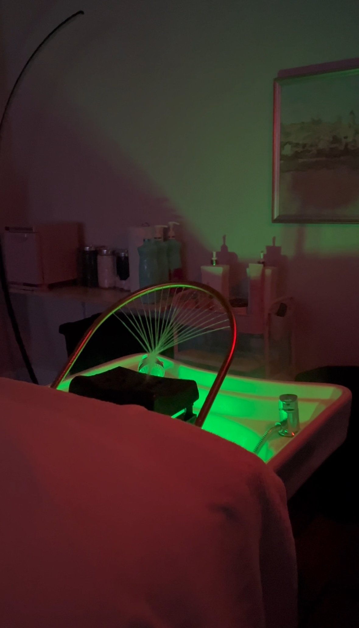 A dark room with an illuminated green-lit head spa with golden halo and a painting on the wall. In the background, there are bottles of shampoos and conditioners in this spokane headspa.
