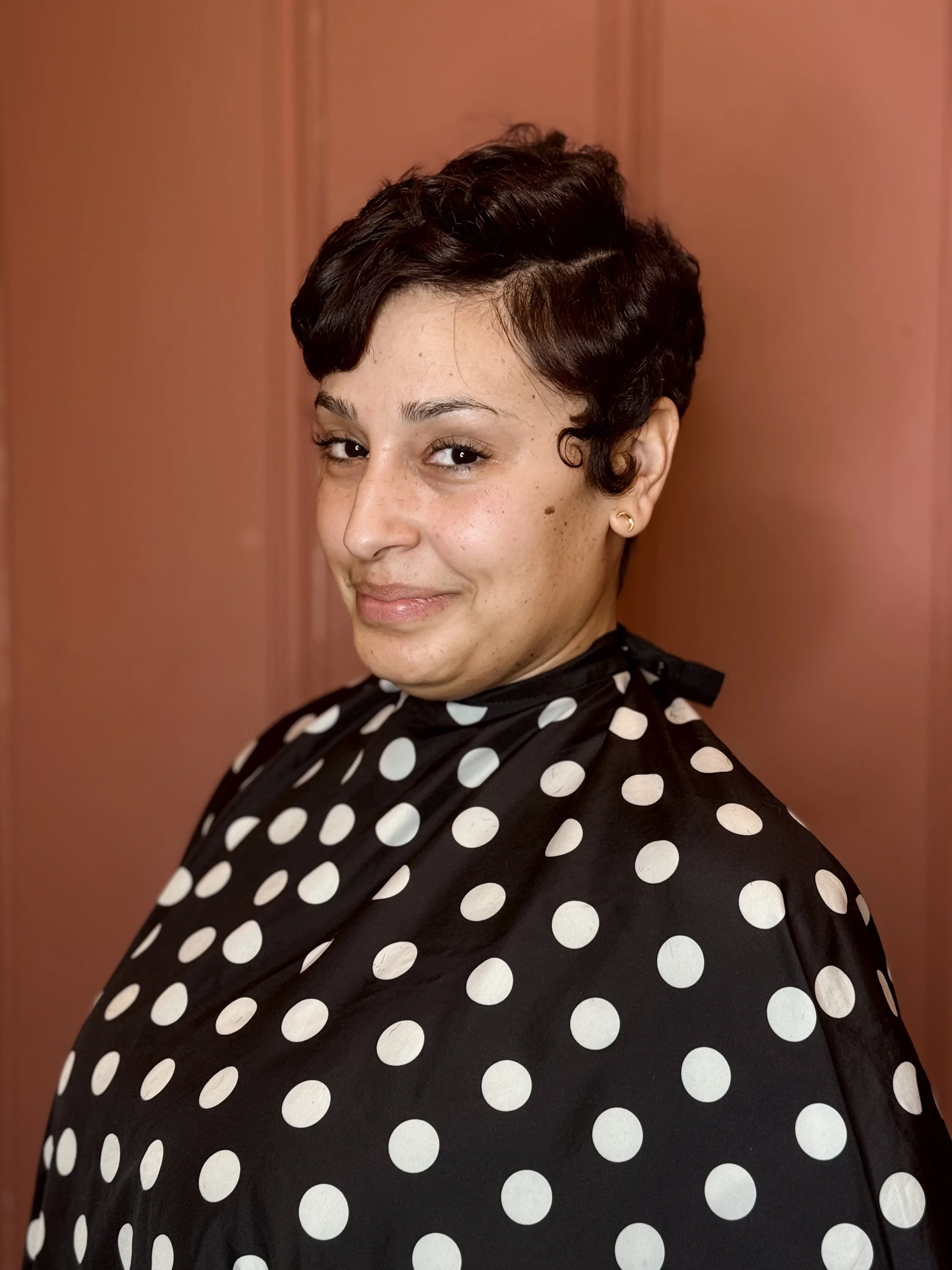 A woman with short dark hair and earrings, wearing a black and white polka dot top, smiling and looking slightly to the side, standing against a pinkish background.