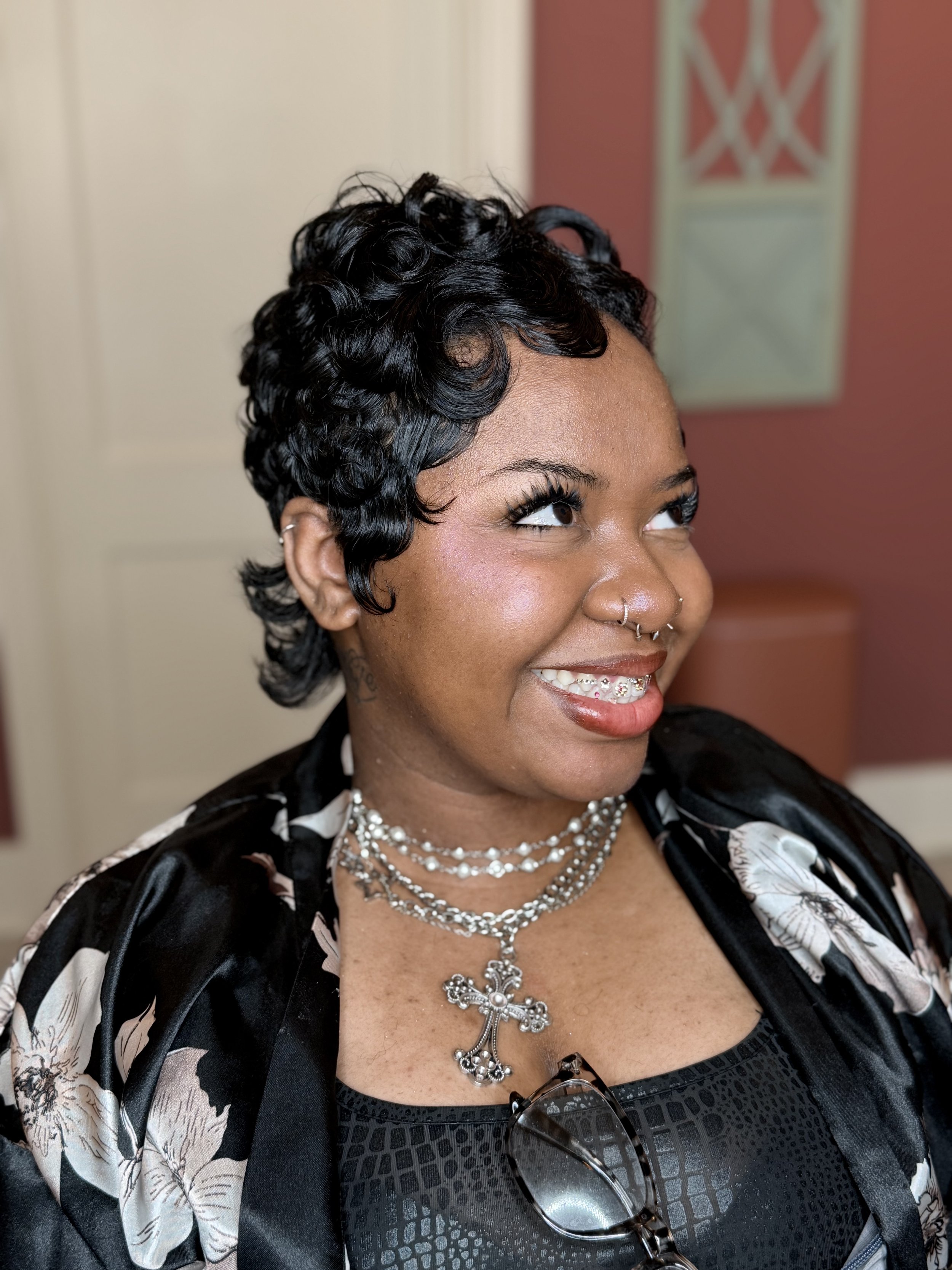A woman with short, curly black hair and multiple layered necklaces, including a large cross pendant, smiling and looking to her left in an indoor setting.