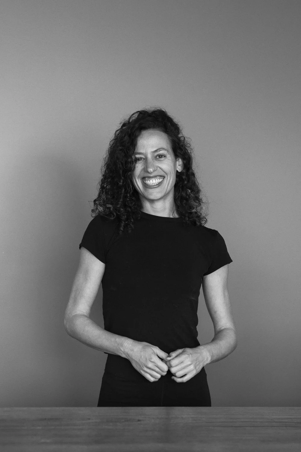 Black-and-white photo of a smiling woman with curly hair, standing against a plain background, wearing a short-sleeve black shirt.