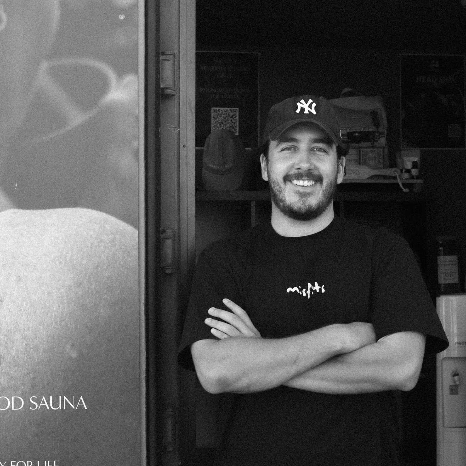 A smiling man wearing a New York Yankees cap and a black t-shirt with white text, standing with arms crossed in front of a dark background with shelves and a QR code. The photo is in black and white.
