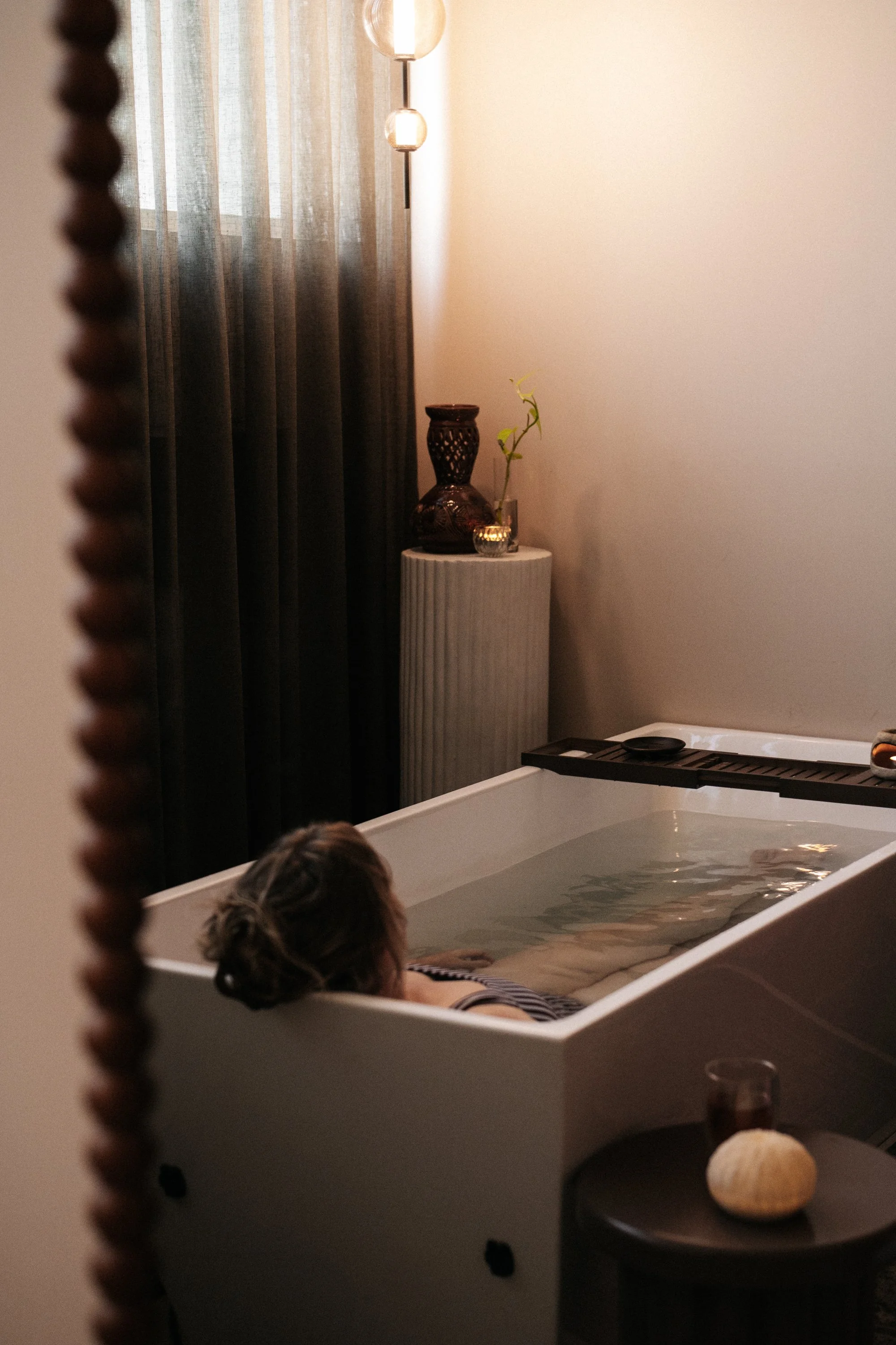 Person relaxing in a bathtub filled with water in a spa or wellness room. The room has a soft, warm lighting with a modern decor, including a white pedestal with decorative vases and a small plant. Curtains cover the window, contributing to a serene atmosphere.
