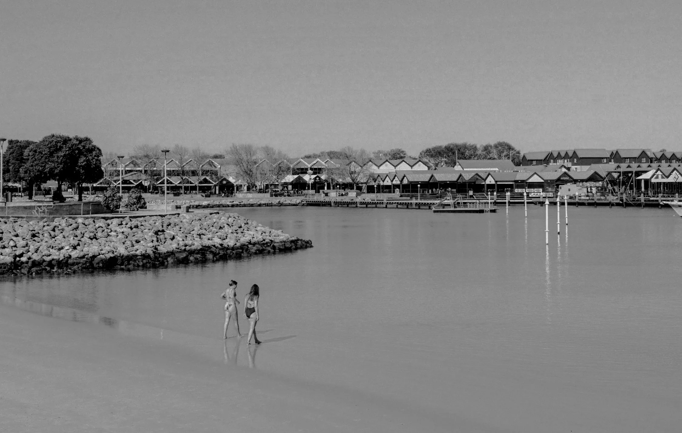 Two women in swimsuits walking along the shallow shoreline of a body of water, with a rocky area nearby and a town with houses and trees in the background.