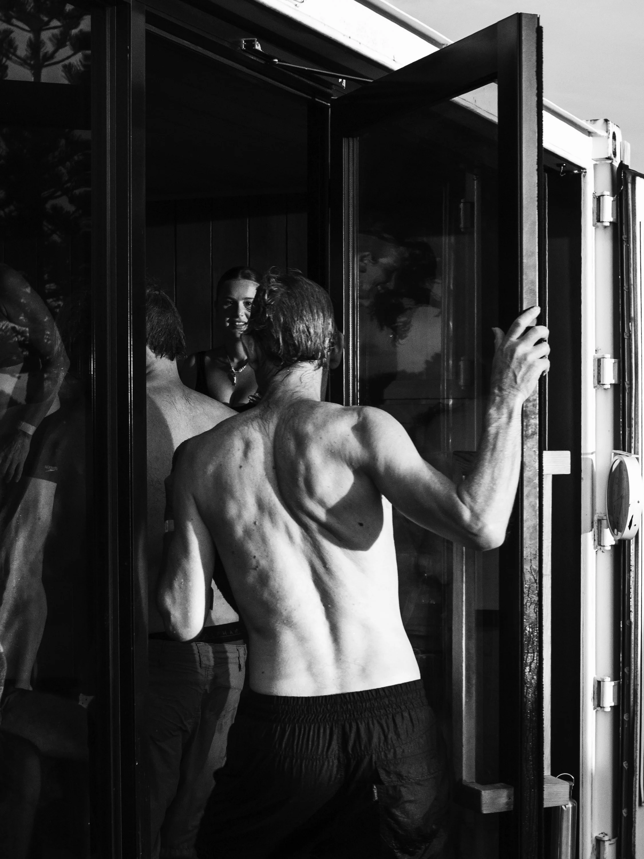 A group of people, including shirtless men and women, are gathered around a glass door, engaging in conversation outdoors.