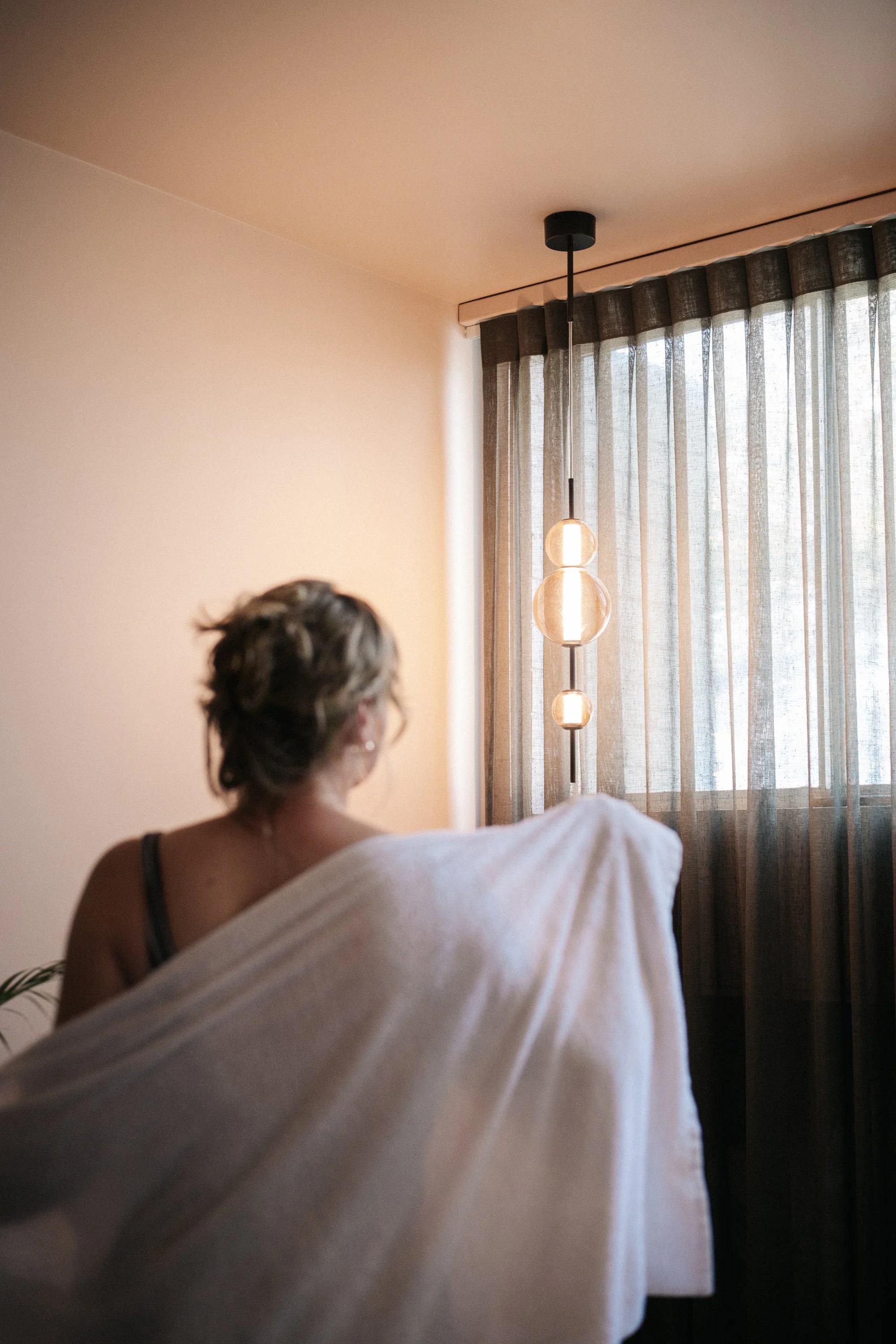 A woman with short hair sitting in a room with sheer curtains, a modern hanging light fixture, and soft natural lighting.