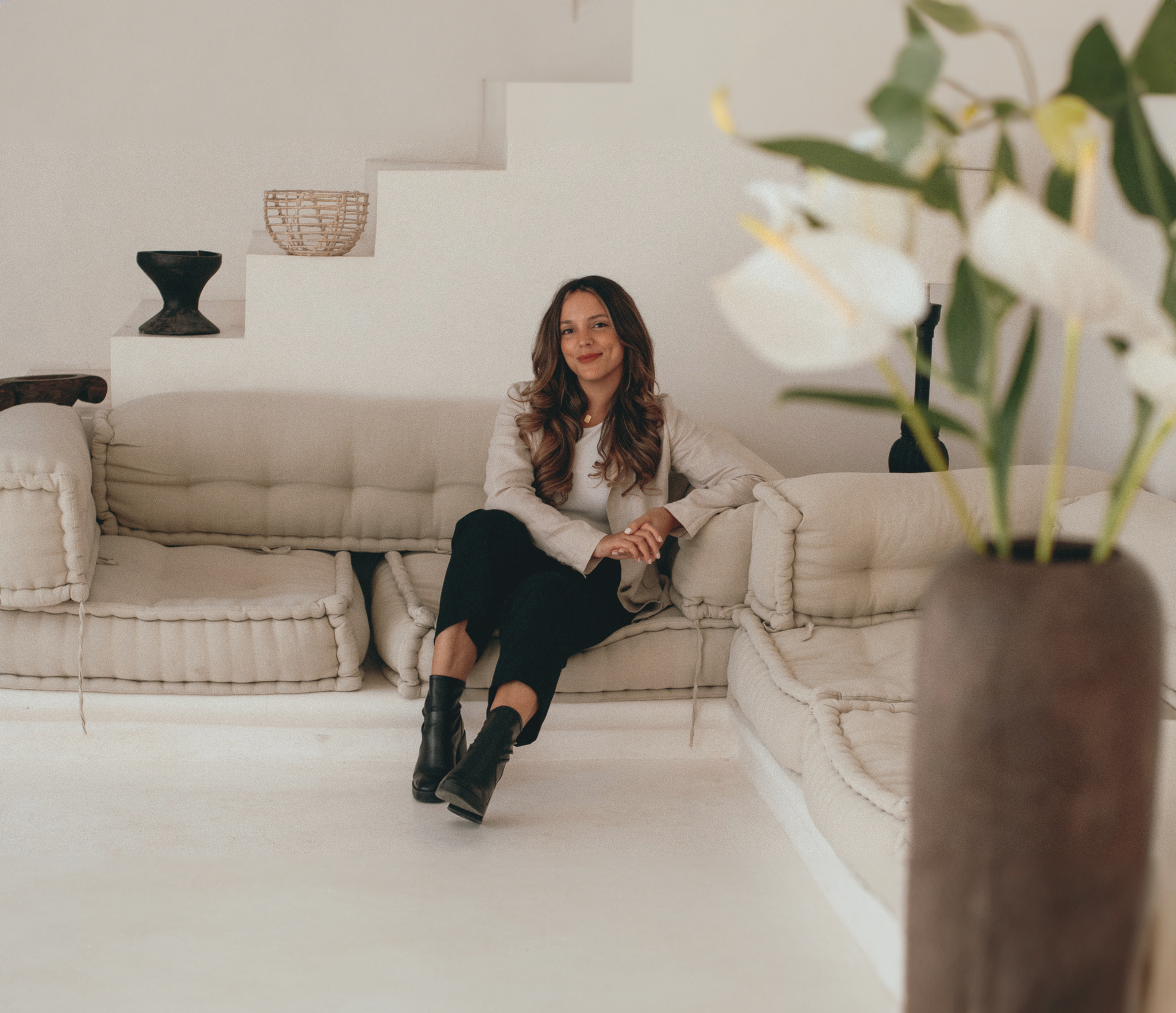 Woman sitting on a modern beige sofa in a minimalist living room with potted plants in the foreground.