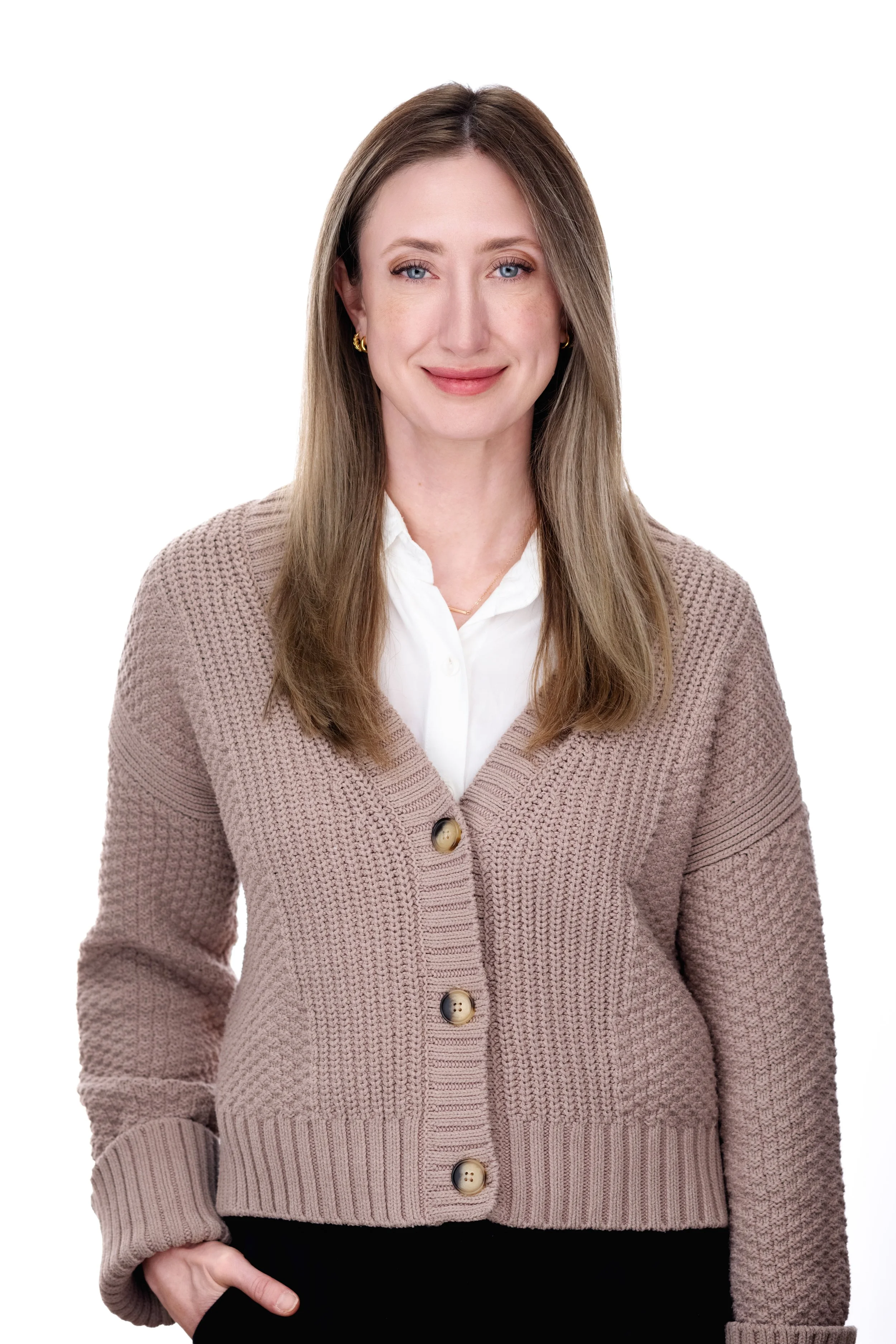 A woman with long brown hair, blue eyes, and gold earrings, wearing a beige knit cardigan over a white shirt, standing against a white background.