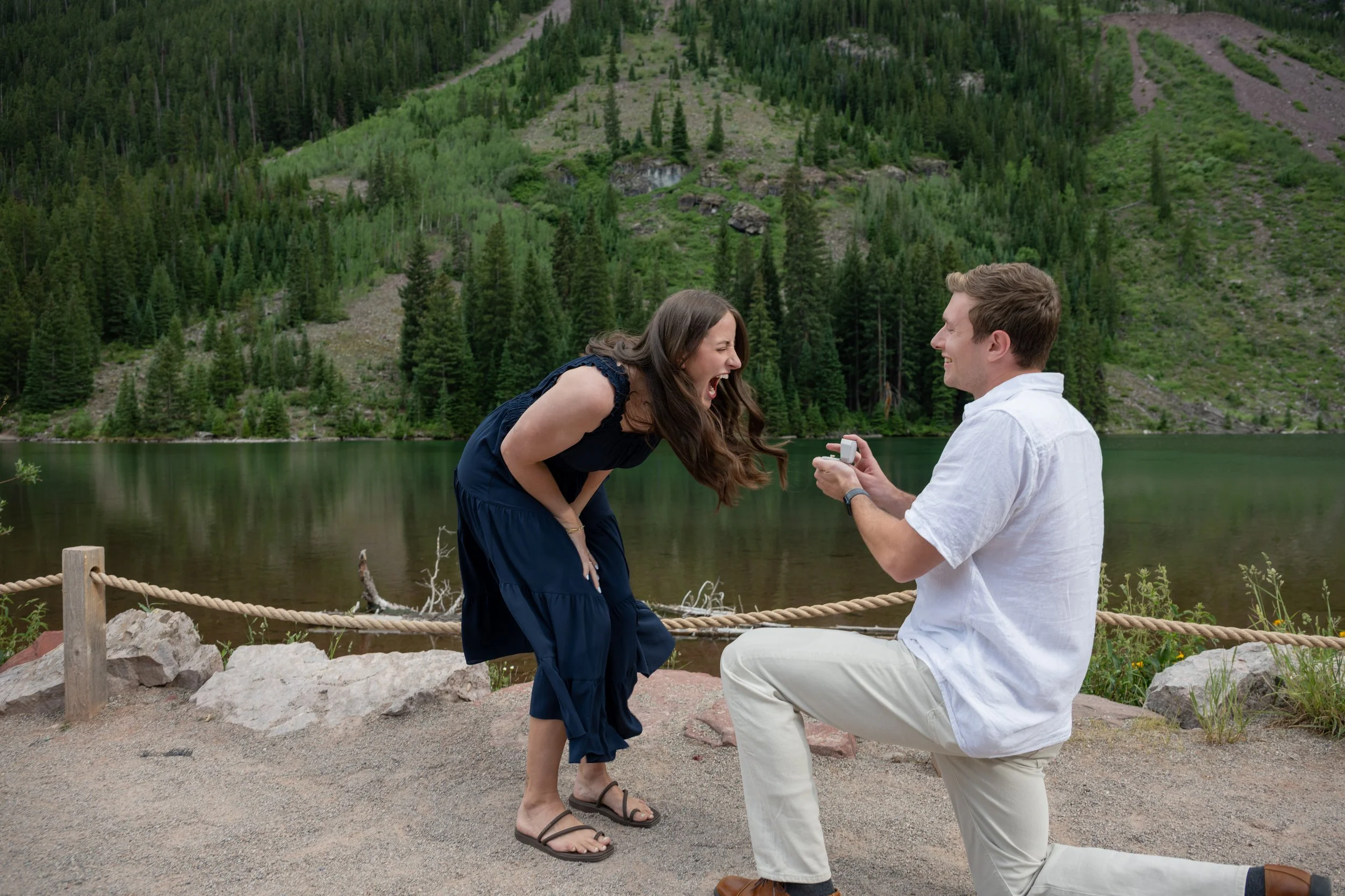 A man on one knee proposing marriage to a woman near a lake surrounded by green trees and mountain landscape, with the woman laughing and leaning forward.