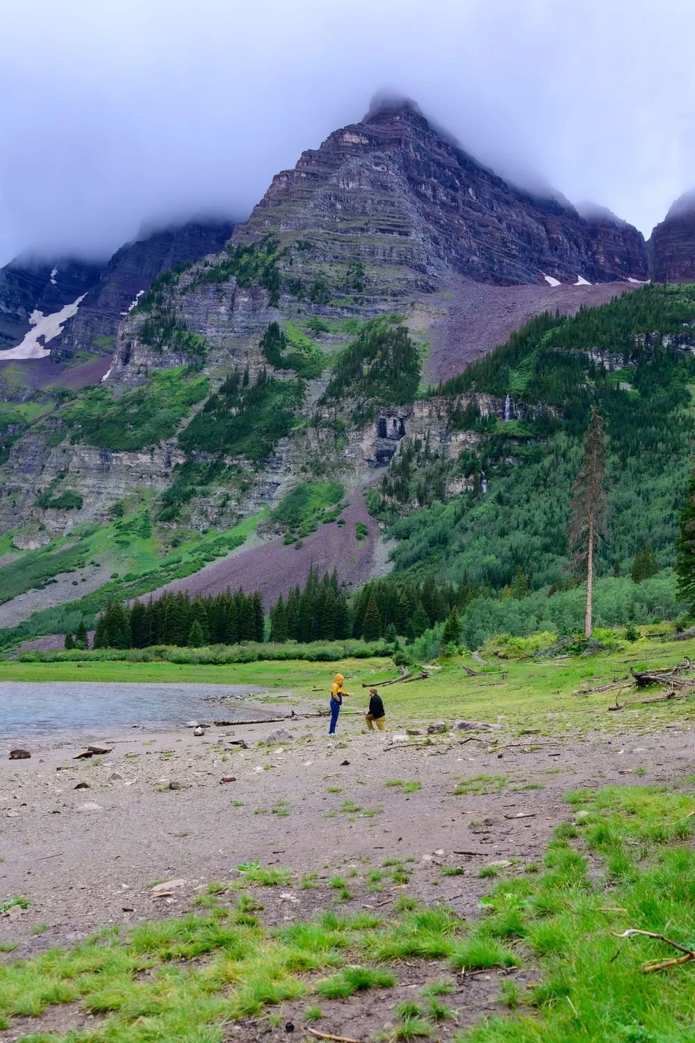 Two people at a lake with a mountainous backdrop, partially covered by clouds, surrounded by greenery and rocky terrain.