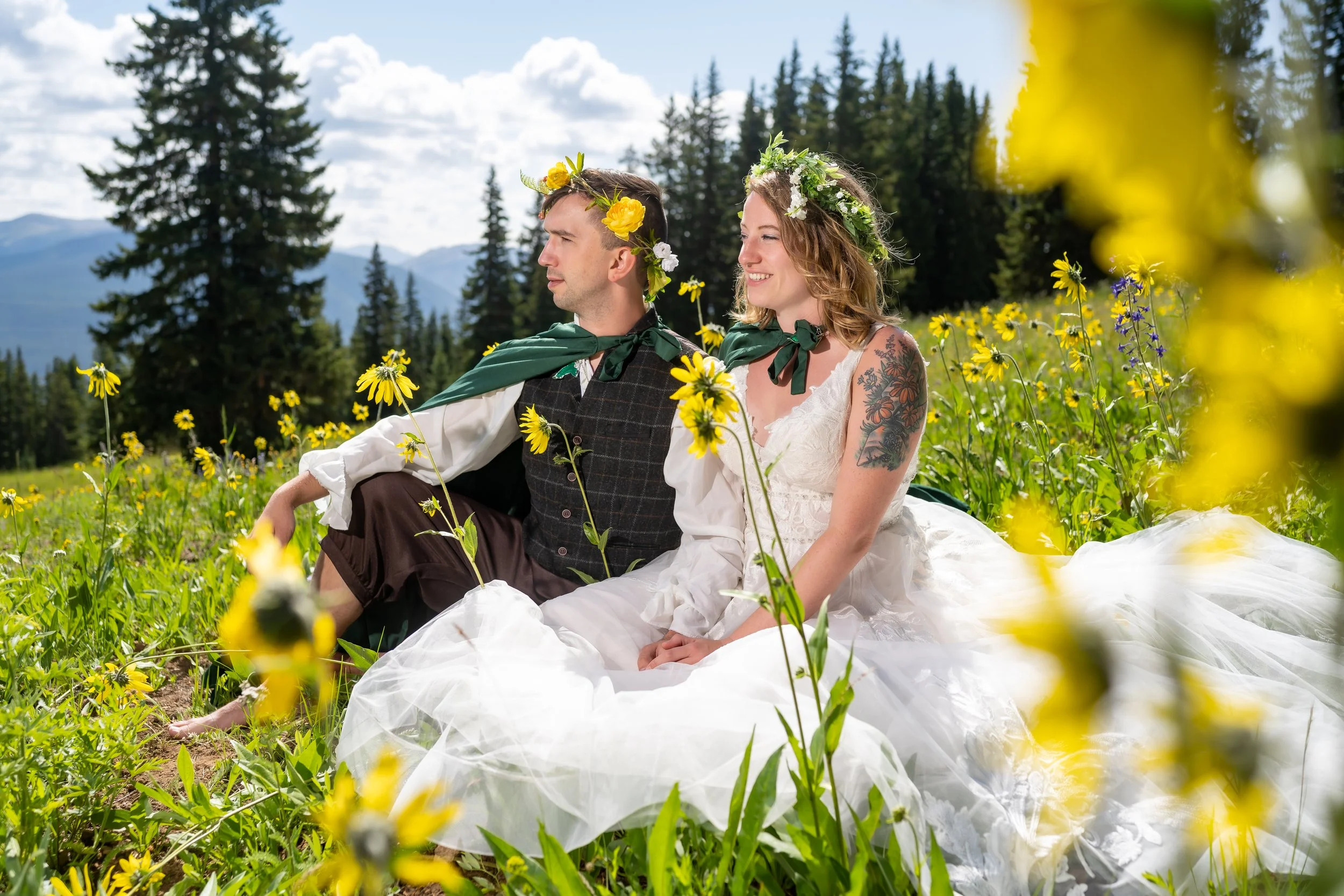 A man and woman dressed as a bride and groom sitting in a field of yellow flowers with pine trees and mountains in the background, wearing floral crowns and green scarves.