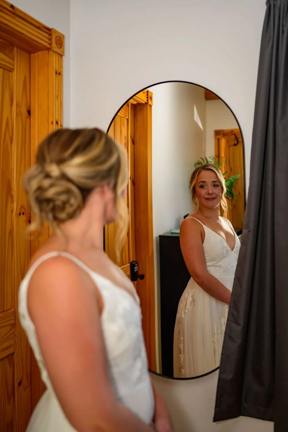 A woman in a white dress looks at herself in an oval mirror, with her hair styled up. She stands in a room with wooden door frames and a gray curtain.