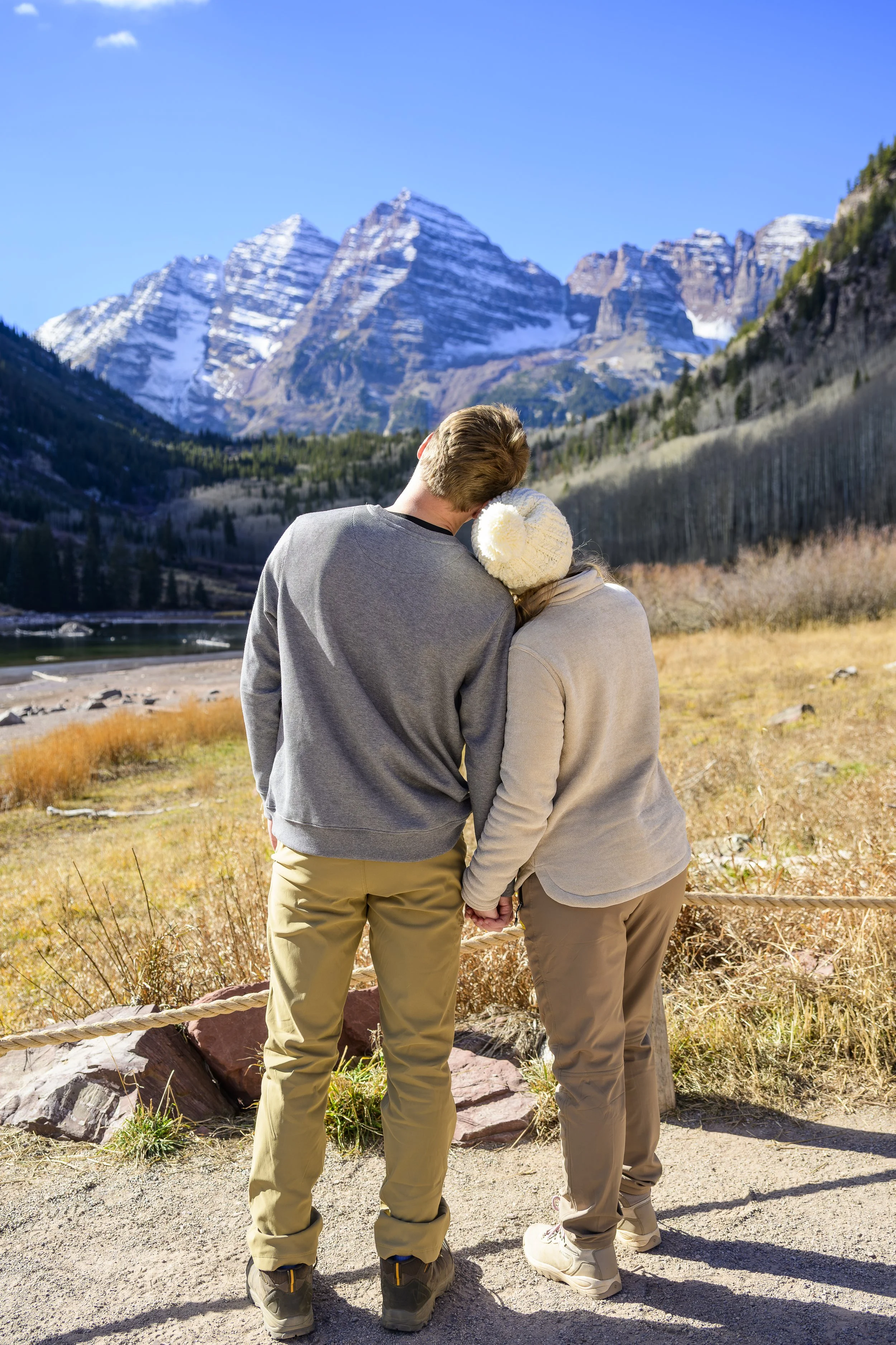 A couple standing together outdoors, holding hands, with snow-capped mountains in the background on a clear, sunny day.