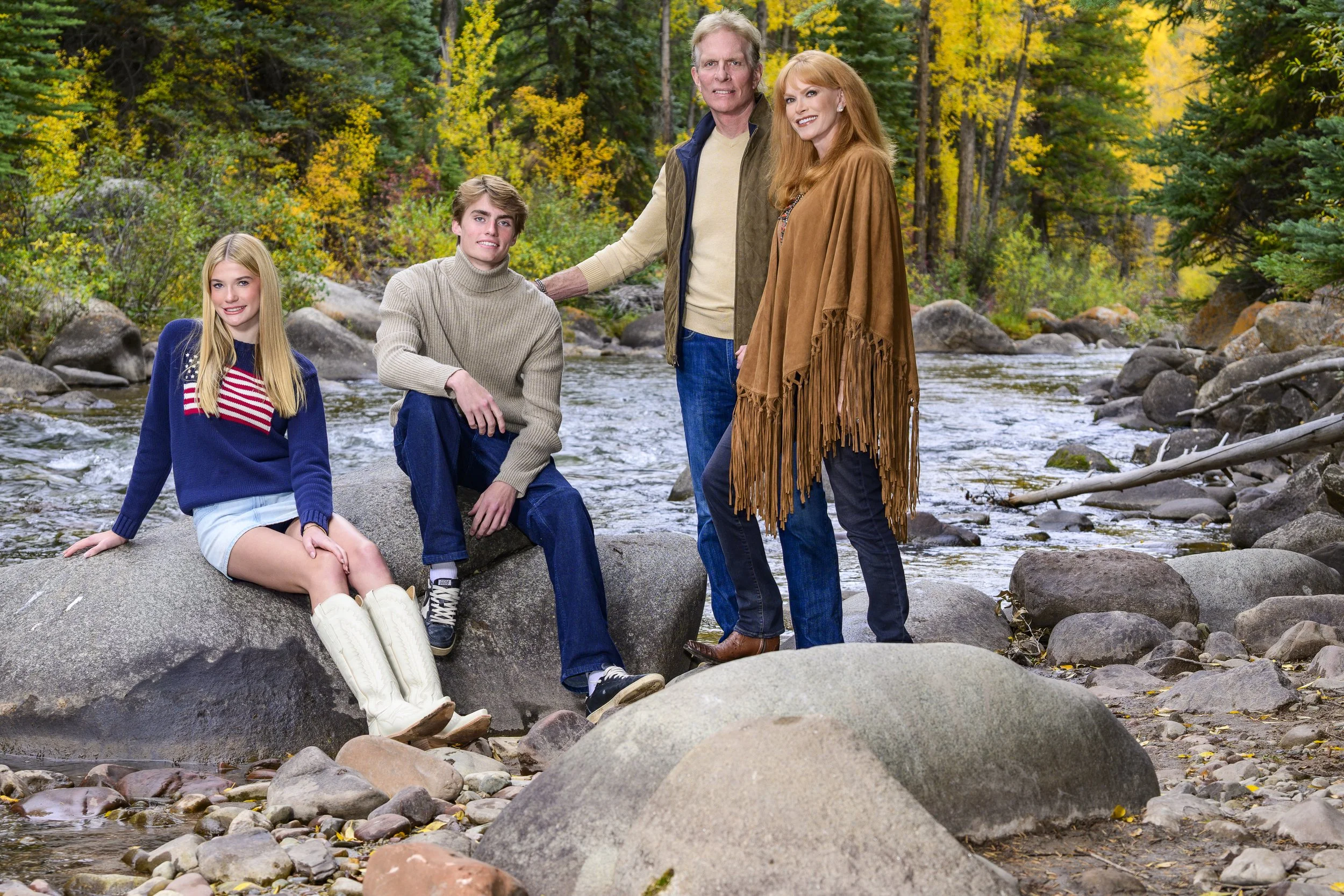 A family of four posing on rocks beside a creek in a forest with autumn foliage.