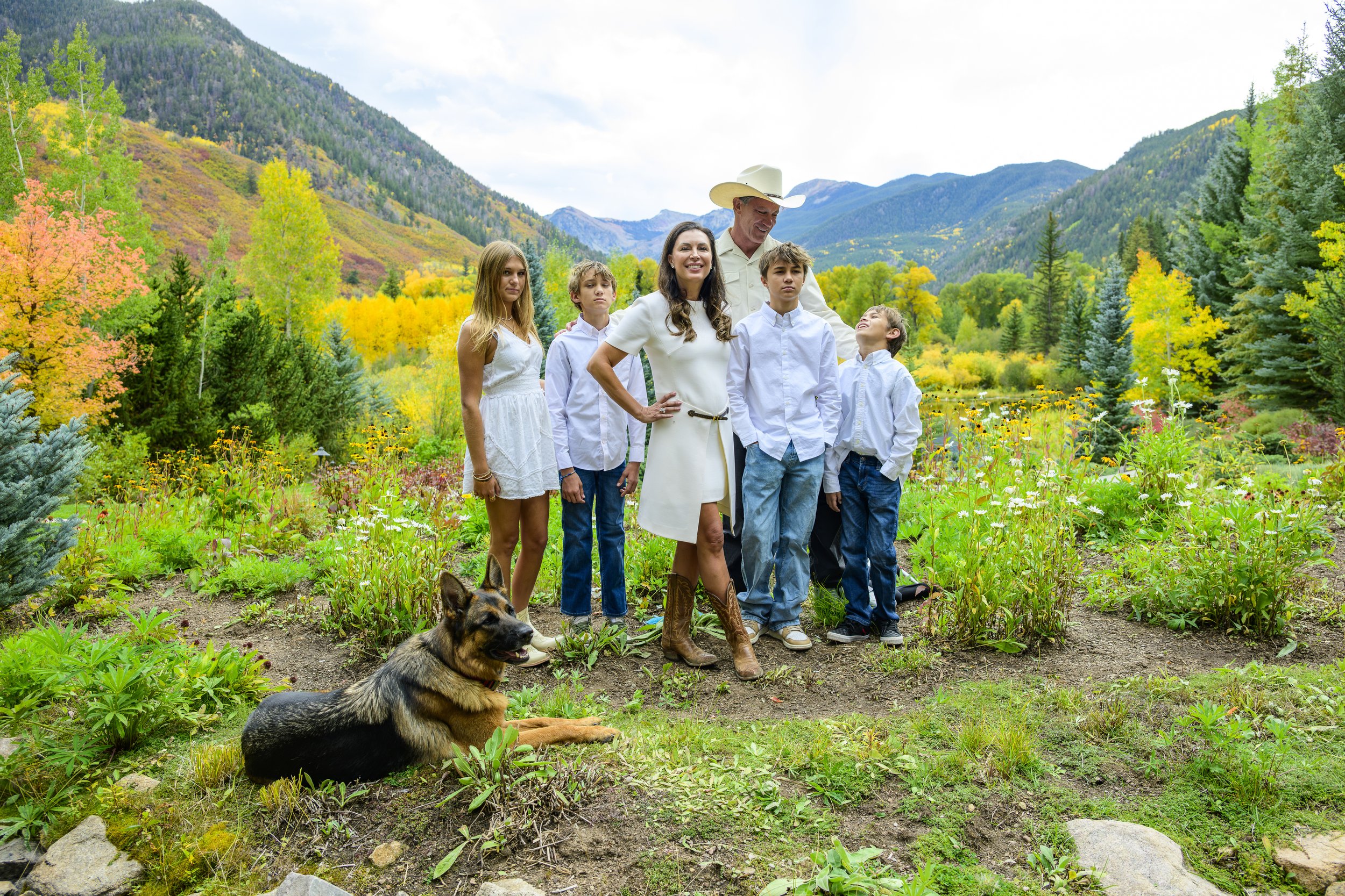 A family of six, including a dog, standing in a lush, colorful garden with mountains in the background.