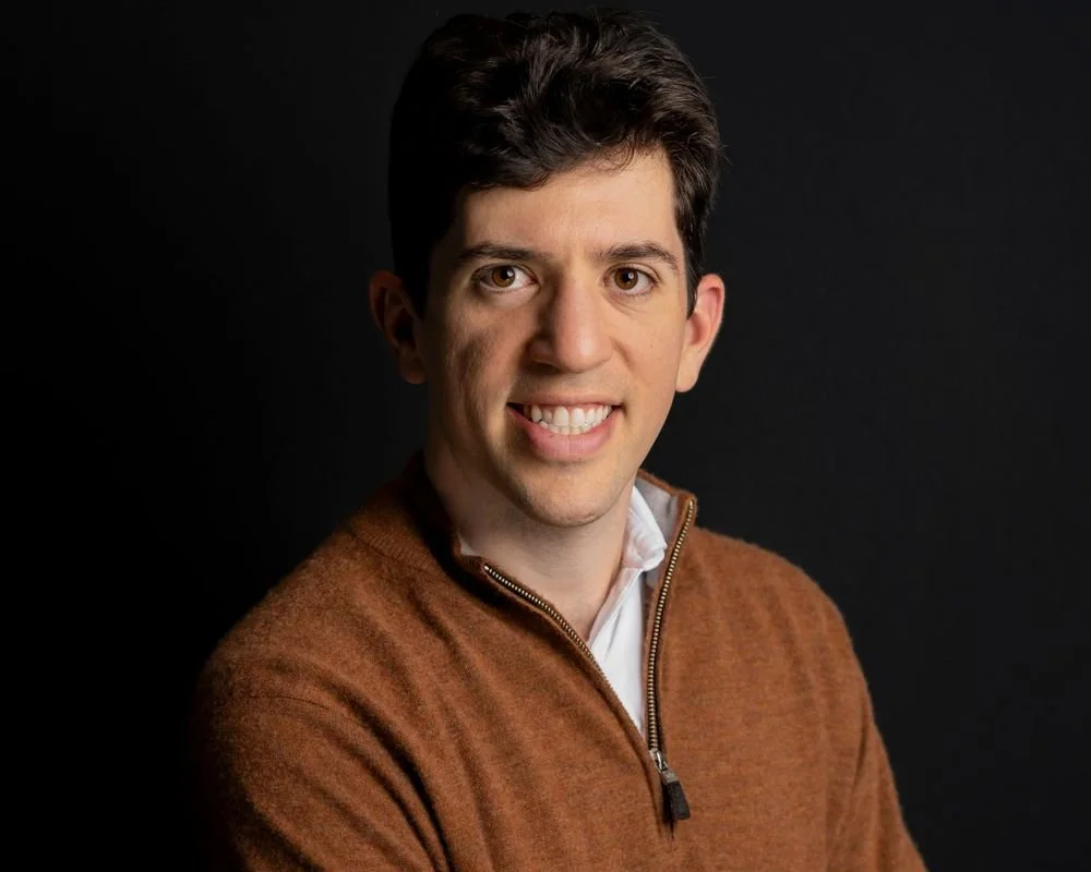 Smiling person in a brown sweater against a black background.