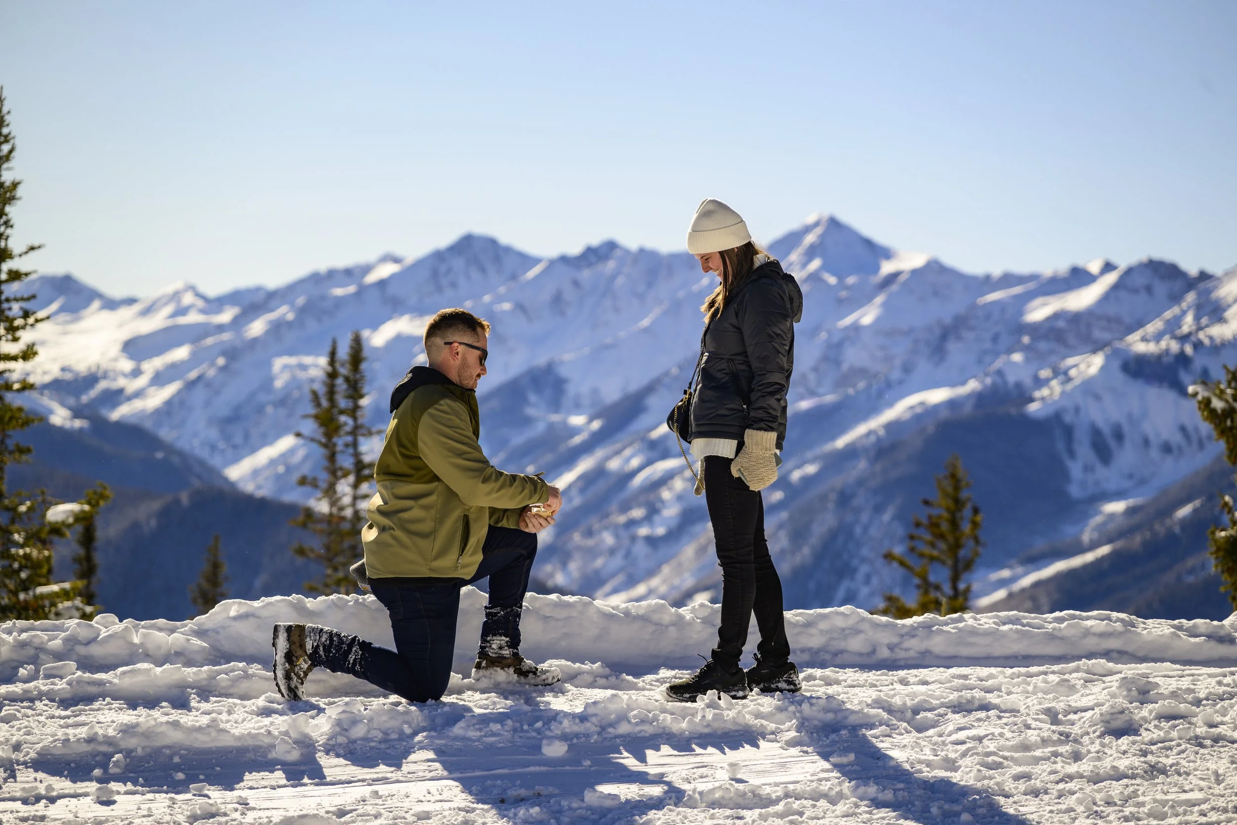 A man proposing to a woman in a snowy mountain landscape with snow-covered peaks and evergreen trees.