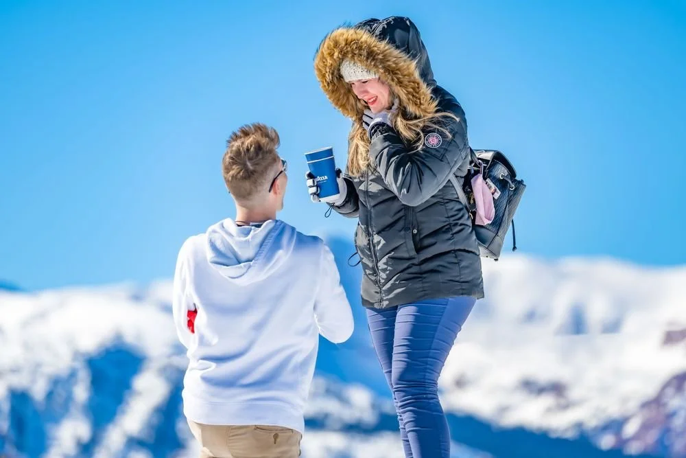 A person in a hoodie kneeling in the snow, proposing to a person in a black winter jacket with a fur hood, holding a cup, against a snowy mountain background.