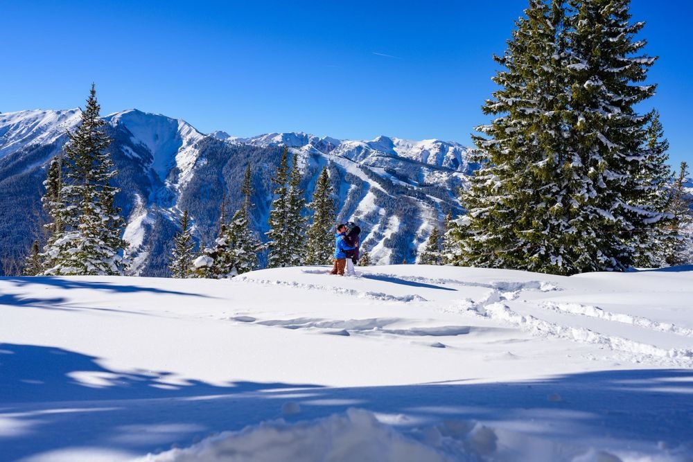 Two people standing in snow-covered landscape with pine trees and snowy mountains in the background on a sunny day.