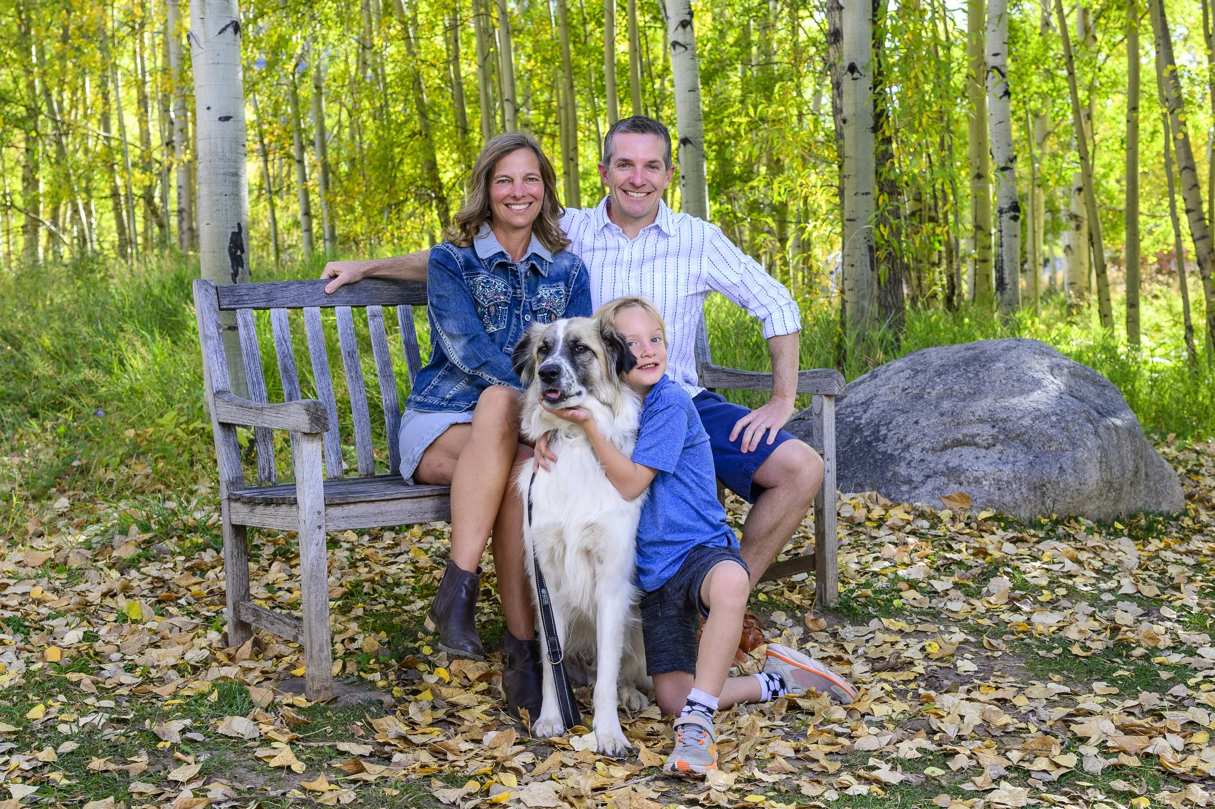 A family of four, with a dog, sitting on a park bench in a forest during autumn, smiling at the camera.