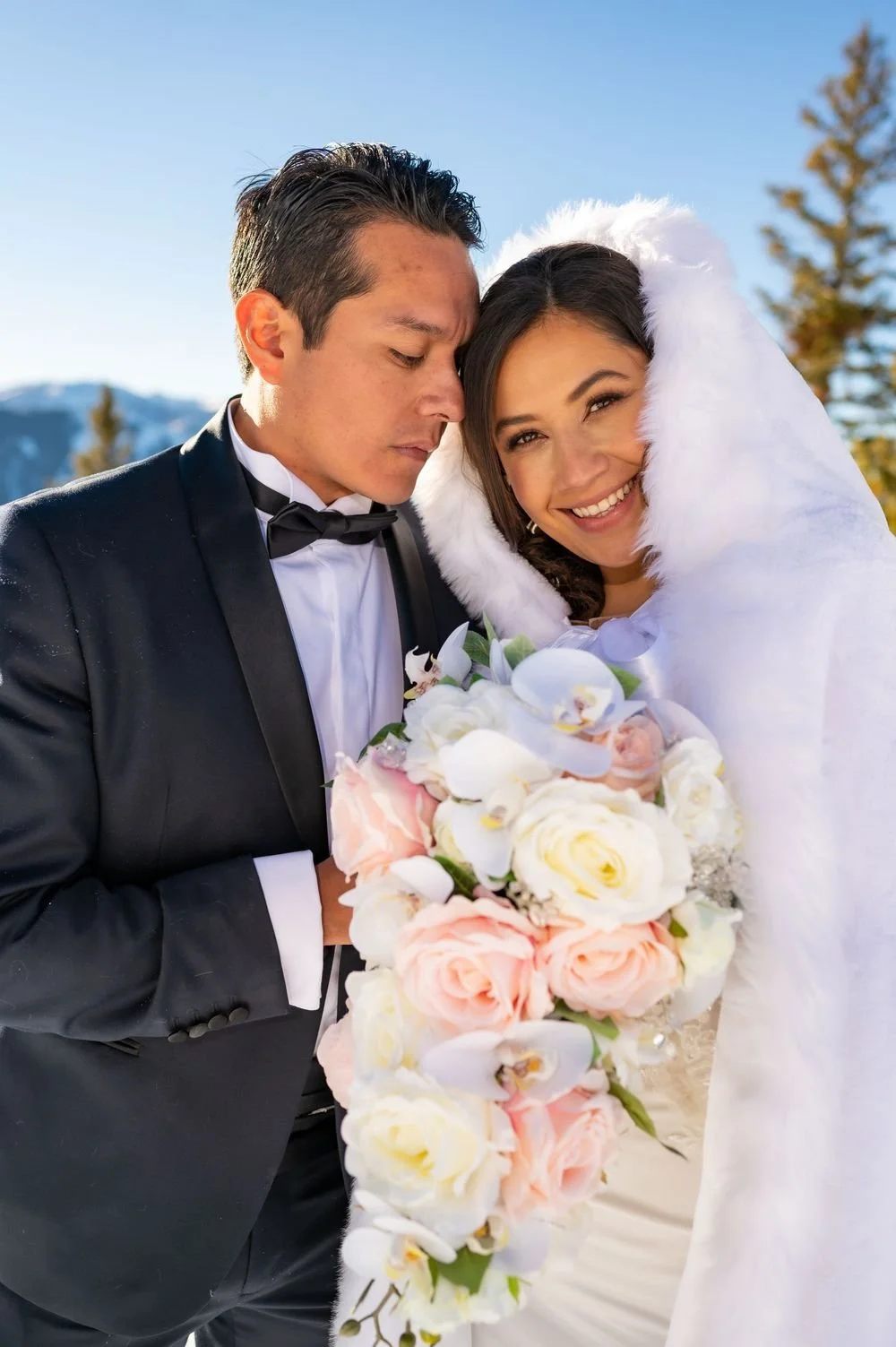 A bride and groom dressed in formal attire, with the bride wearing a white fur hood and holding a bouquet of pink and white flowers, standing outdoors in a snowy landscape.