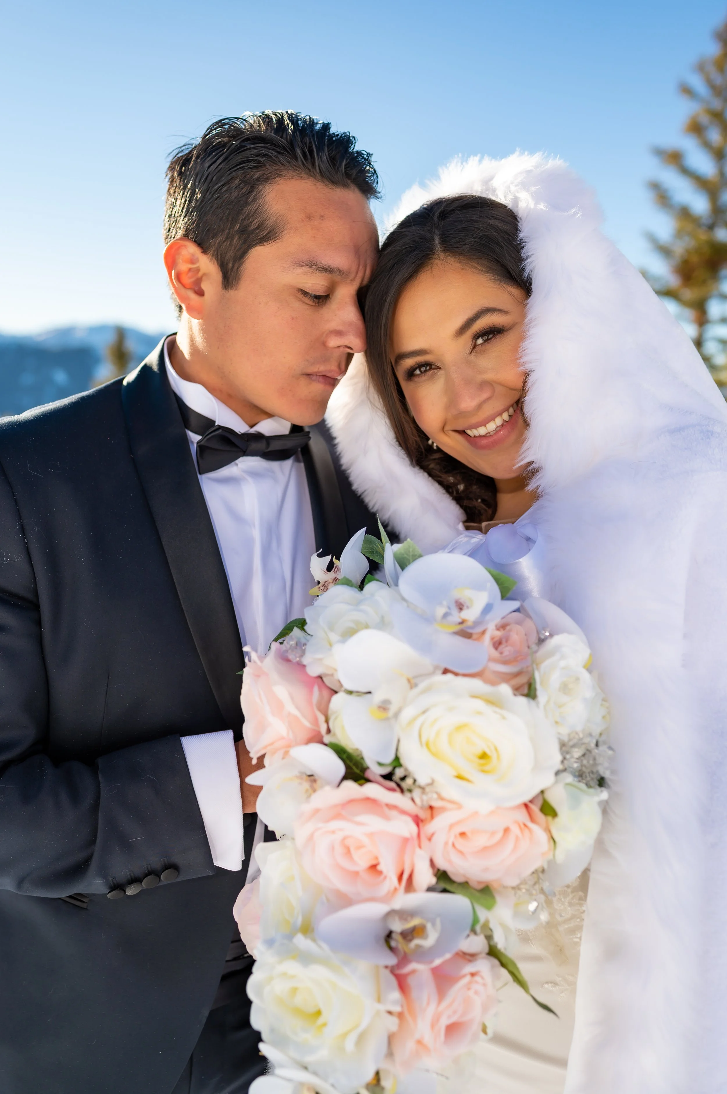 A bride and groom standing outdoors on a sunny day, dressed in wedding attire. The groom is in a black tuxedo with a bow tie, and the bride is smiling with her eyes closed, wrapped in a white fur cover, holding a bouquet of pink, white, and cream-col