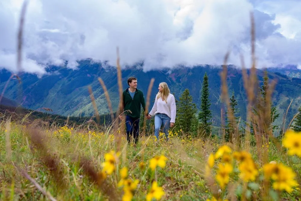 A couple walking hand in hand through a scenic mountain meadow with yellow wildflowers in the foreground and a cloudy sky above.