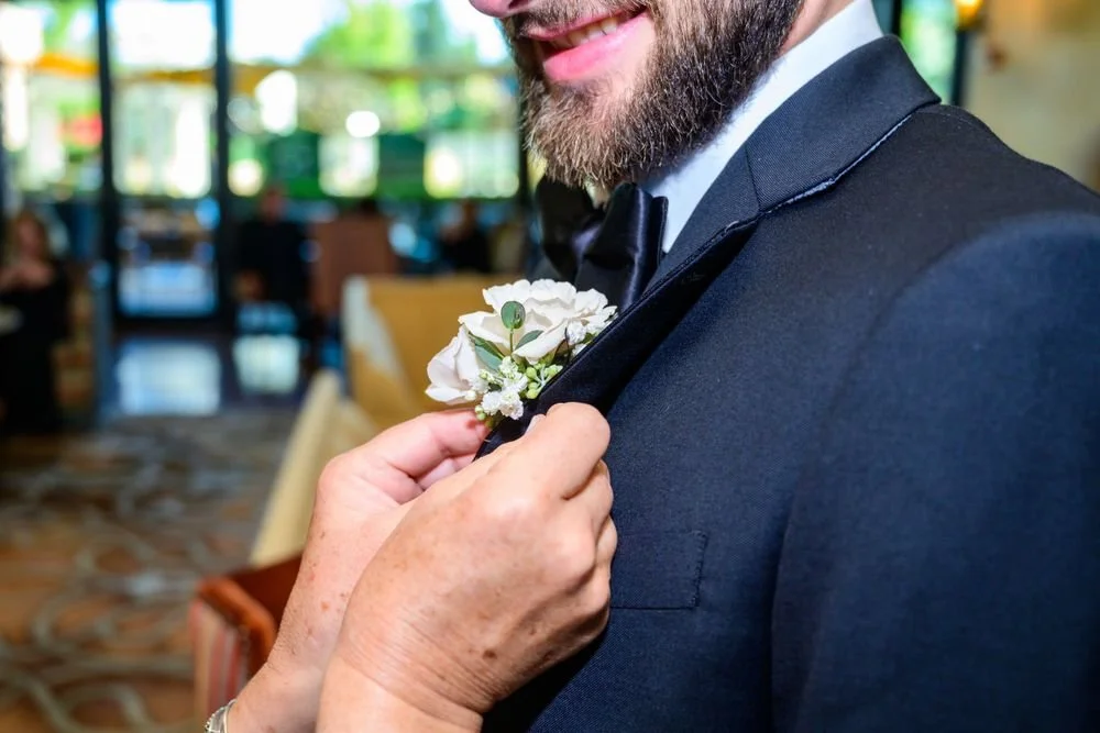 Person in a suit having boutonniere pinned on jacket lapel, indoors, with blurred background.