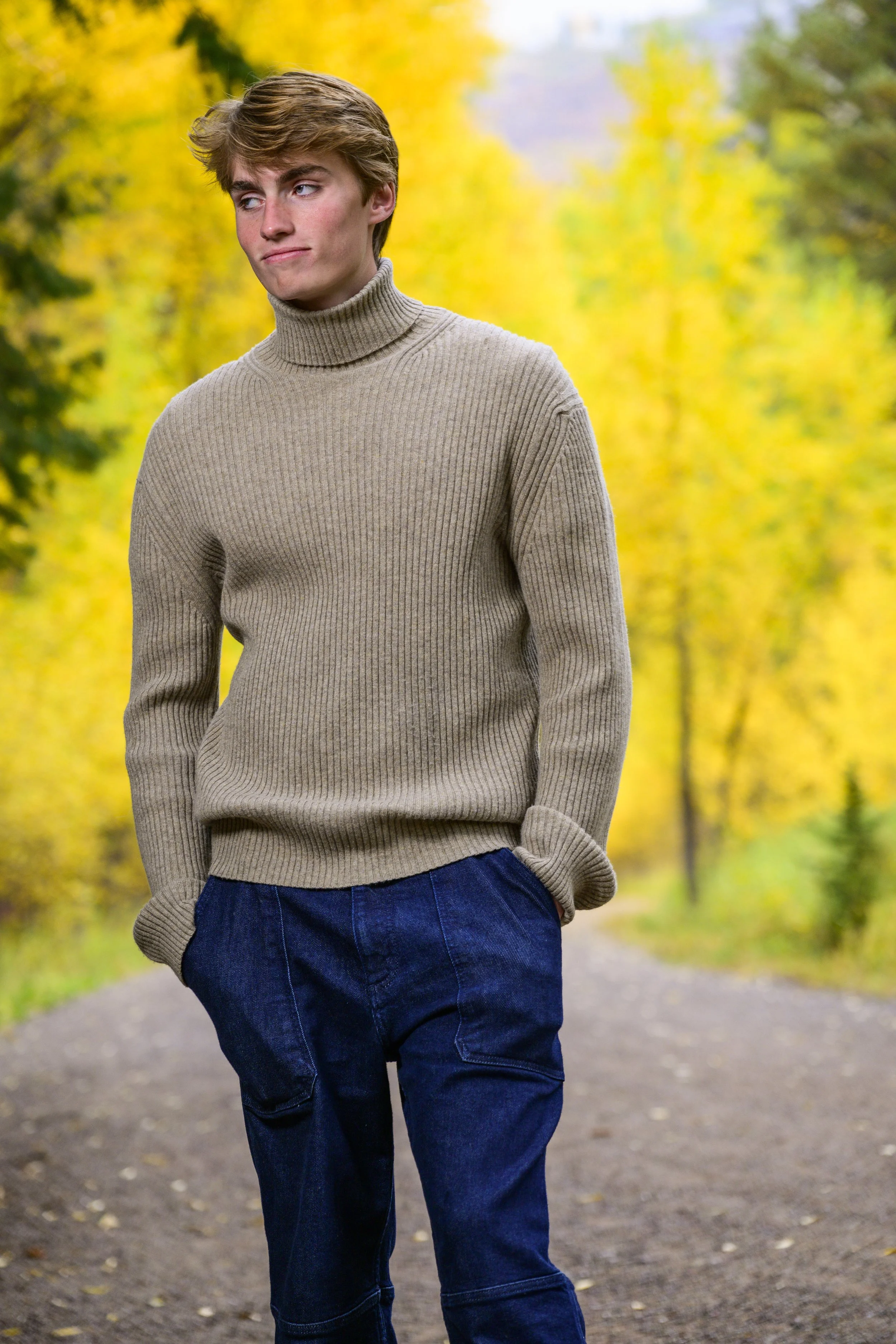 Young man in a beige turtleneck sweater and dark blue jeans standing on a fall-colored forest path with yellow and green foliage.