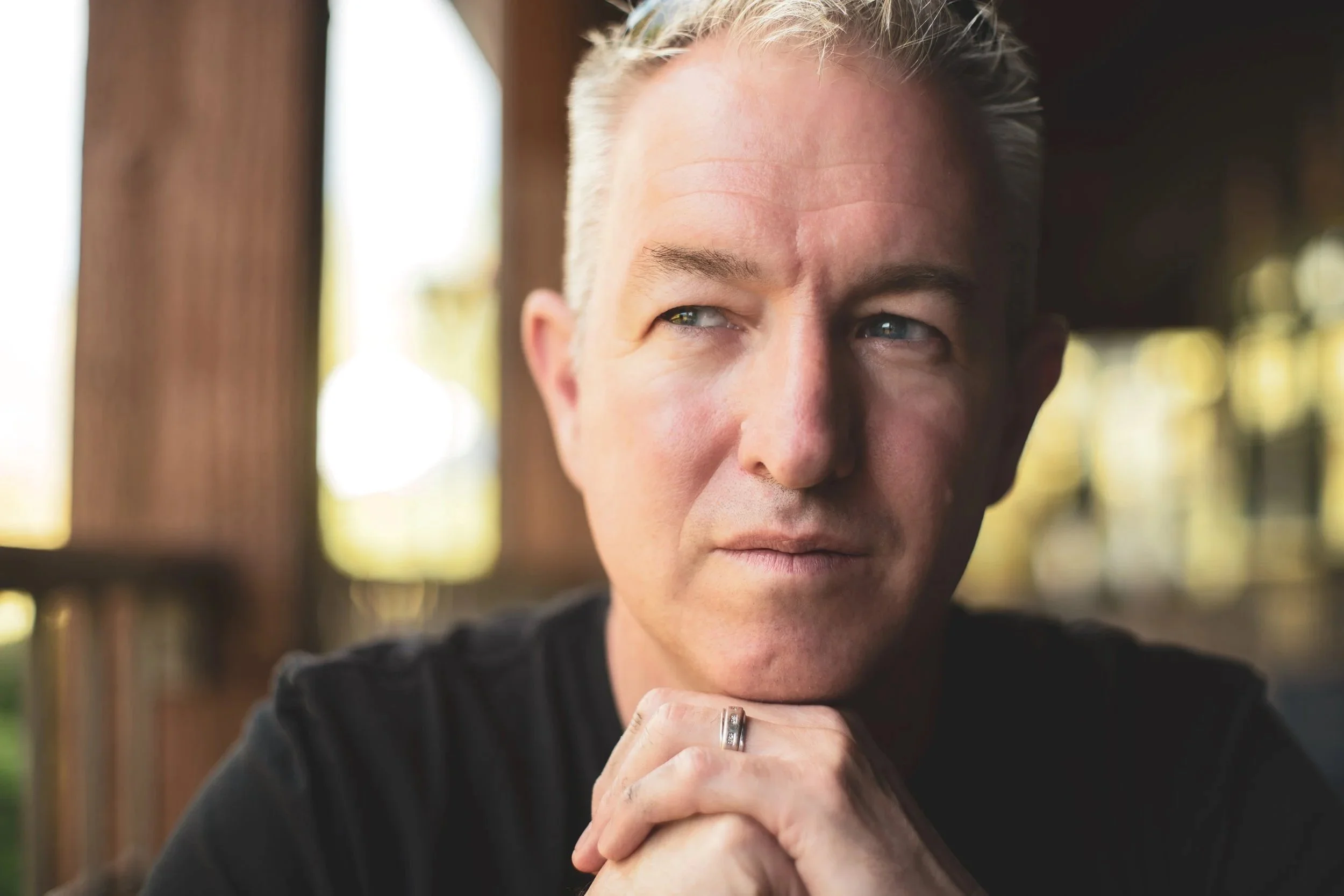 A close-up of a man with blond hair and blue eyes, gazing thoughtfully, with his hand near his chin, wearing a black shirt and a silver ring, standing outdoors with blurred wooden structures and greenery in the background.