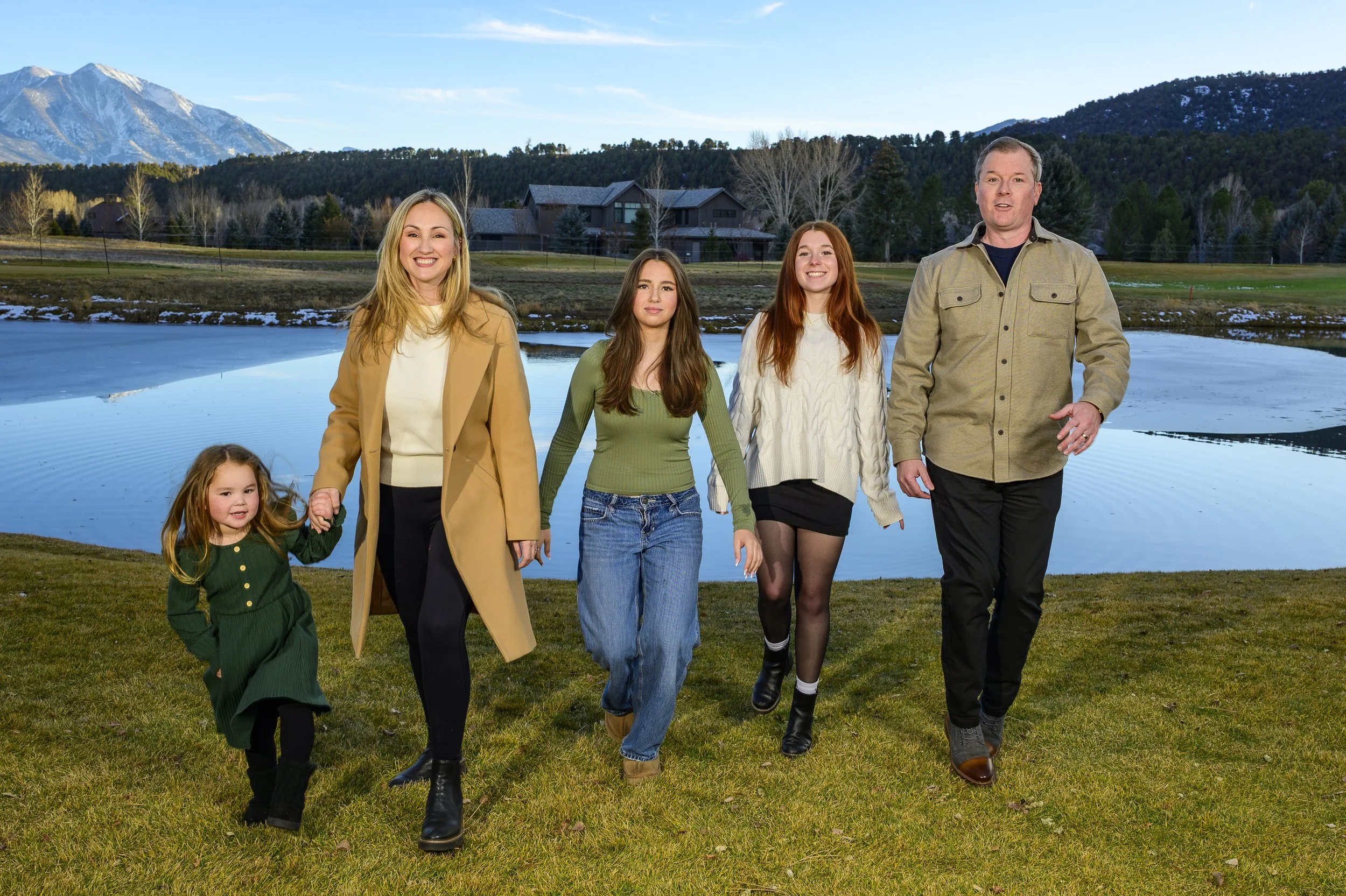 A family of six walking together outdoors near a pond with snow-capped mountains in the background, enjoying a scenic landscape.