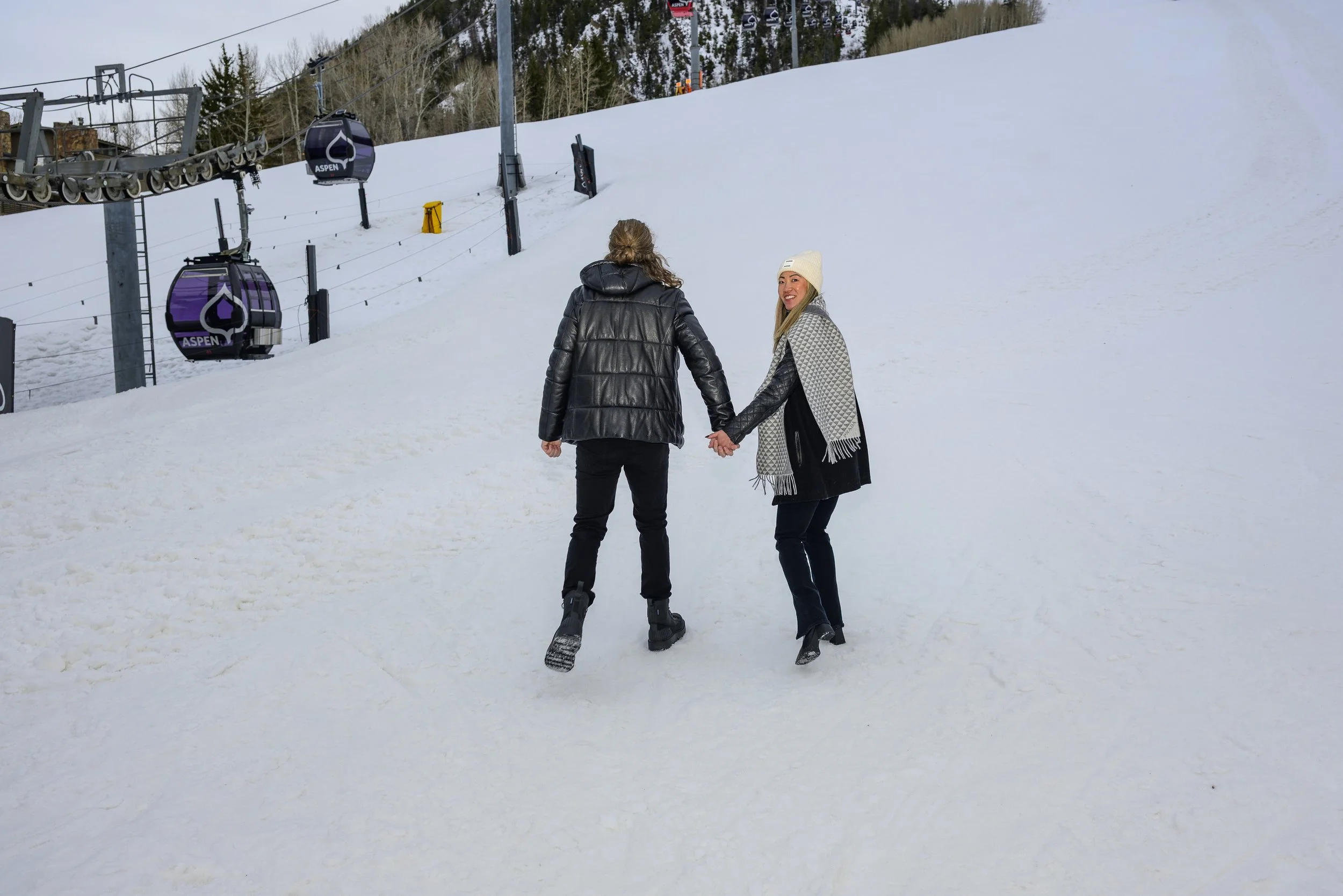 A couple holding hands and walking on a snowy ski slope, with ski lift gondolas in the background at Aspen ski resort.