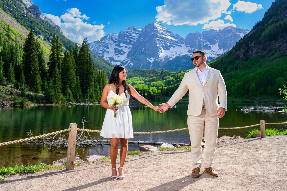 A couple holding hands in front of a scenic mountain lake, surrounded by lush green forests and snow-capped peaks under a bright blue sky.
