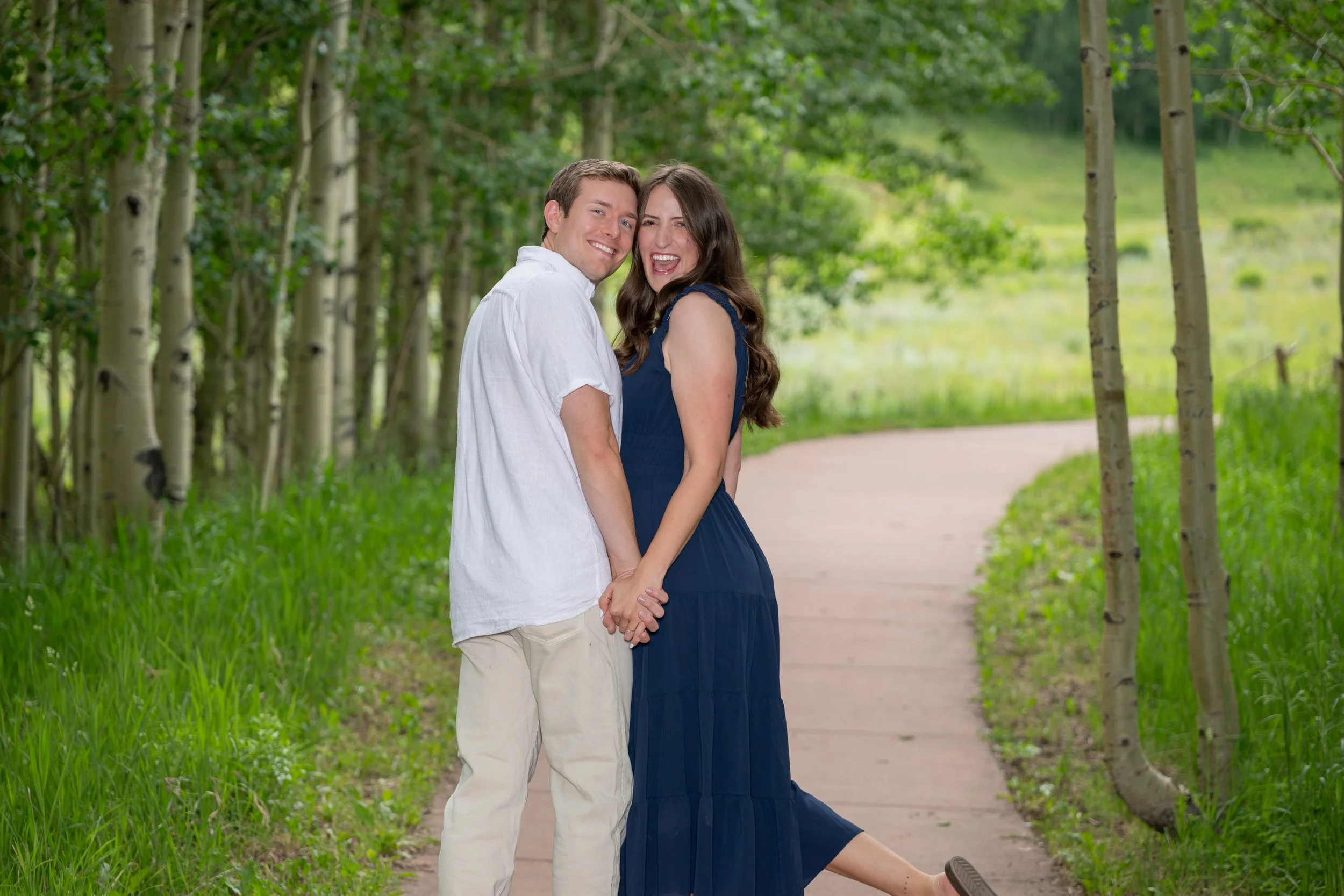 A couple smiling and holding hands on a walking trail in a wooded area.