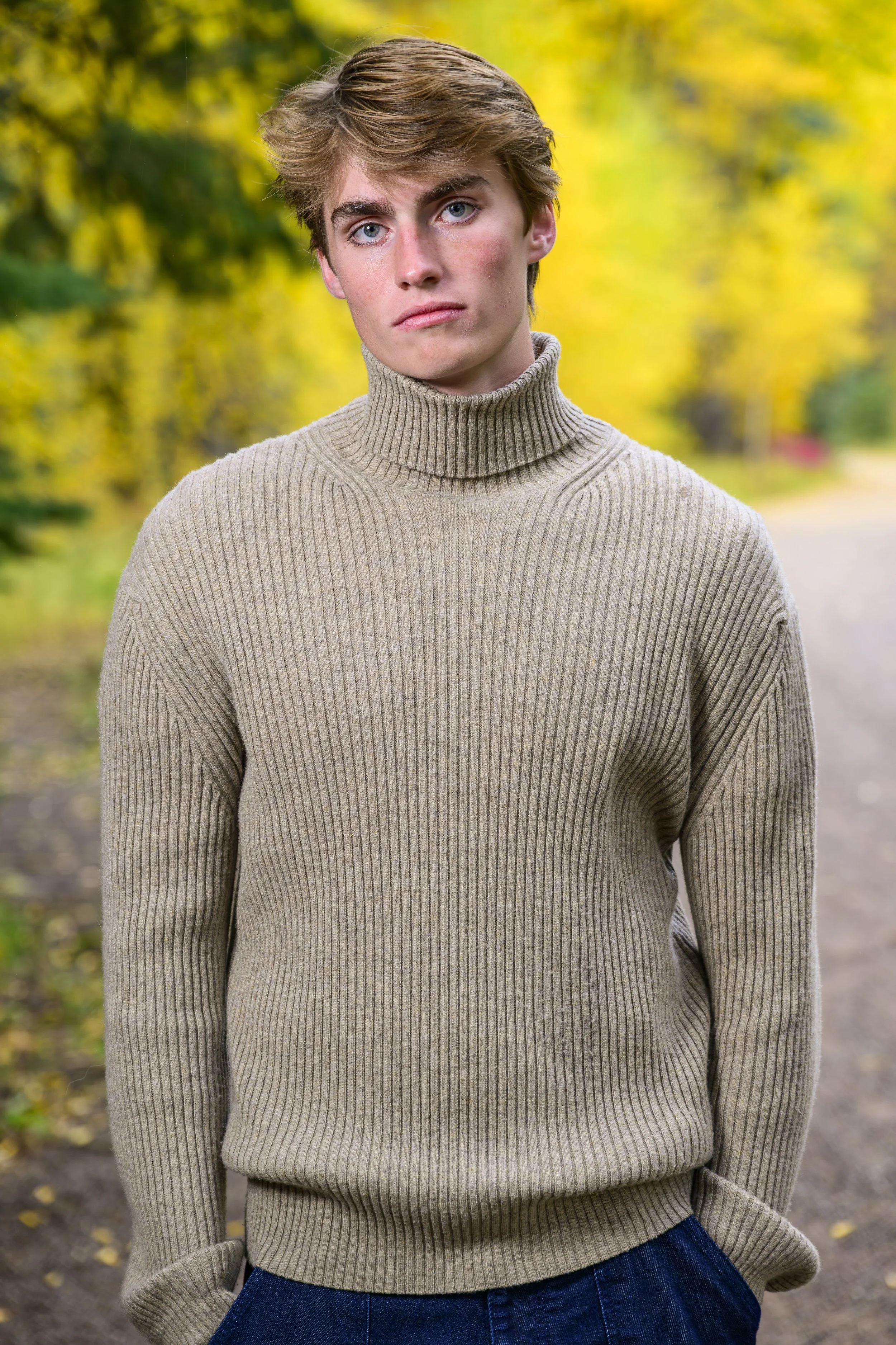 A young man with light brown hair and blue eyes stands outdoors on a dirt path, wearing a beige turtleneck sweater and blue jeans, with autumn trees displaying yellow leaves in the background.