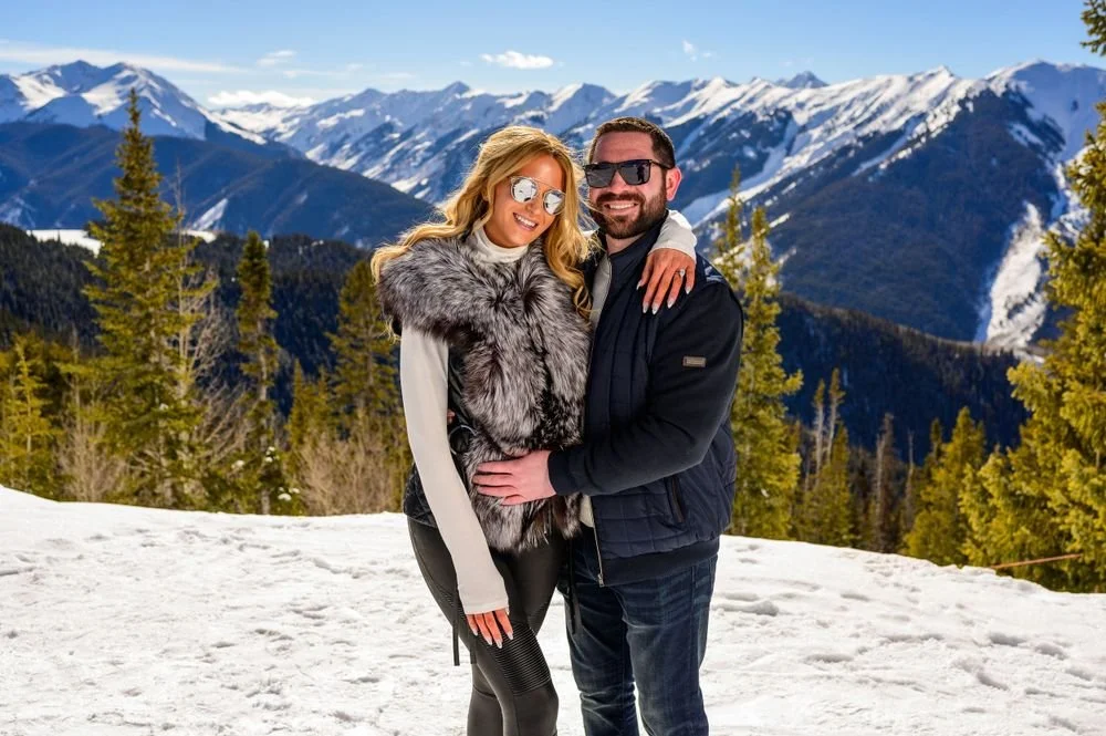 A couple poses on a snowy mountain with scenic views of distant snow-capped peaks and evergreen trees under a clear blue sky.