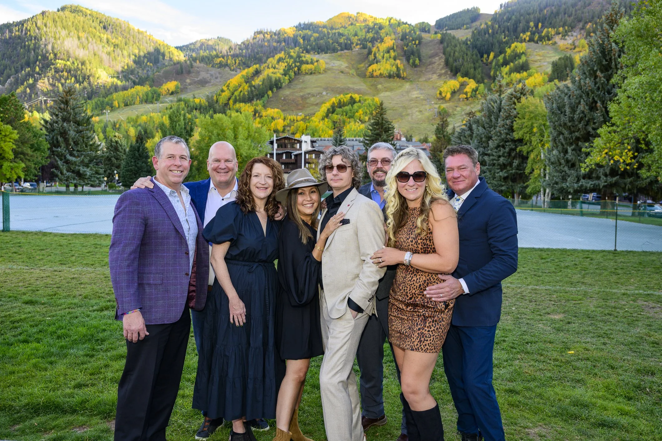Group of nine people smiling and posing together outdoors on a grassy field with trees and mountains in the background, some wearing suits, dresses, and sunglasses.